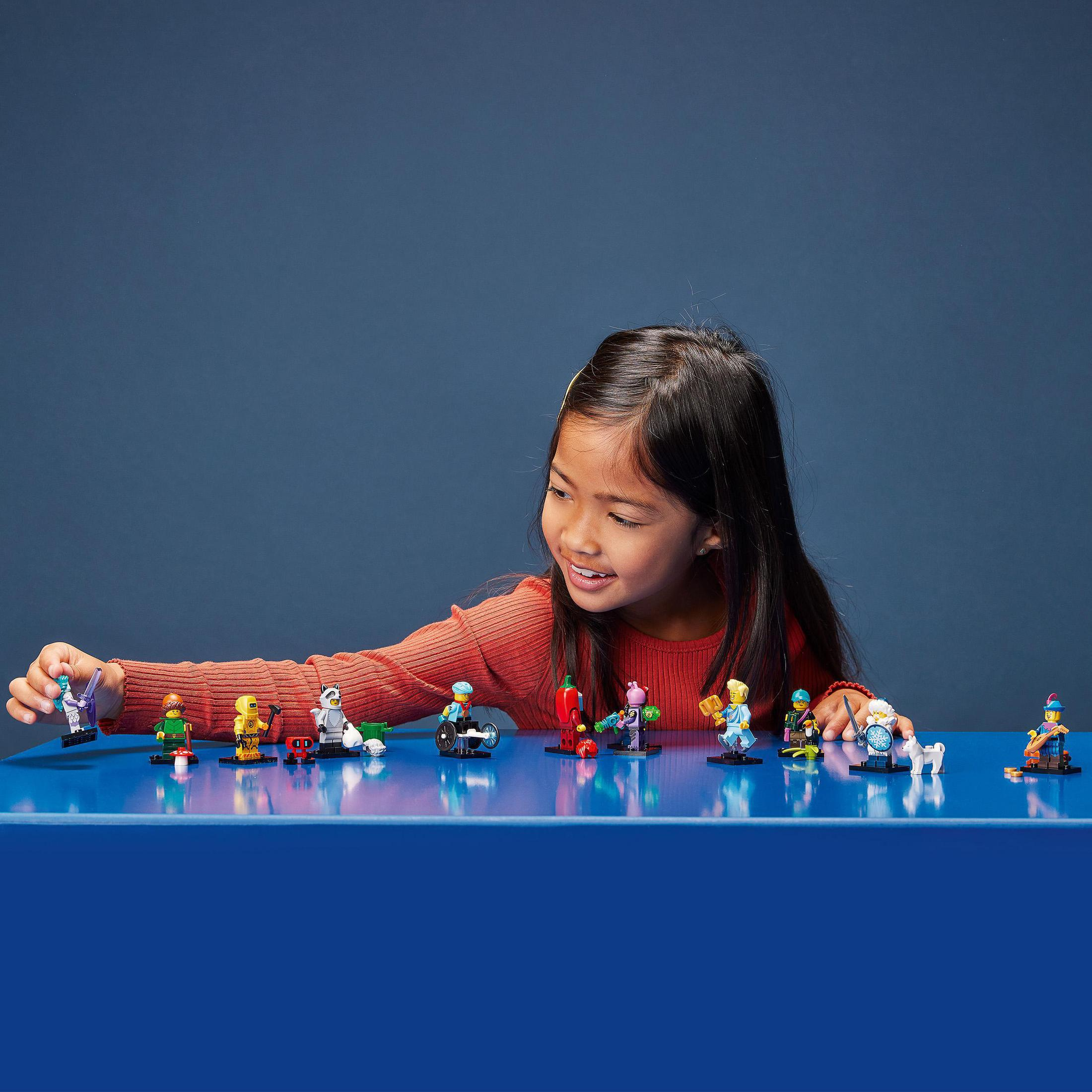 Ein junges Mädchen interagiert mit LEGO-Minifiguren, angeordnet auf einer blauen Oberfläche, lächelnd.