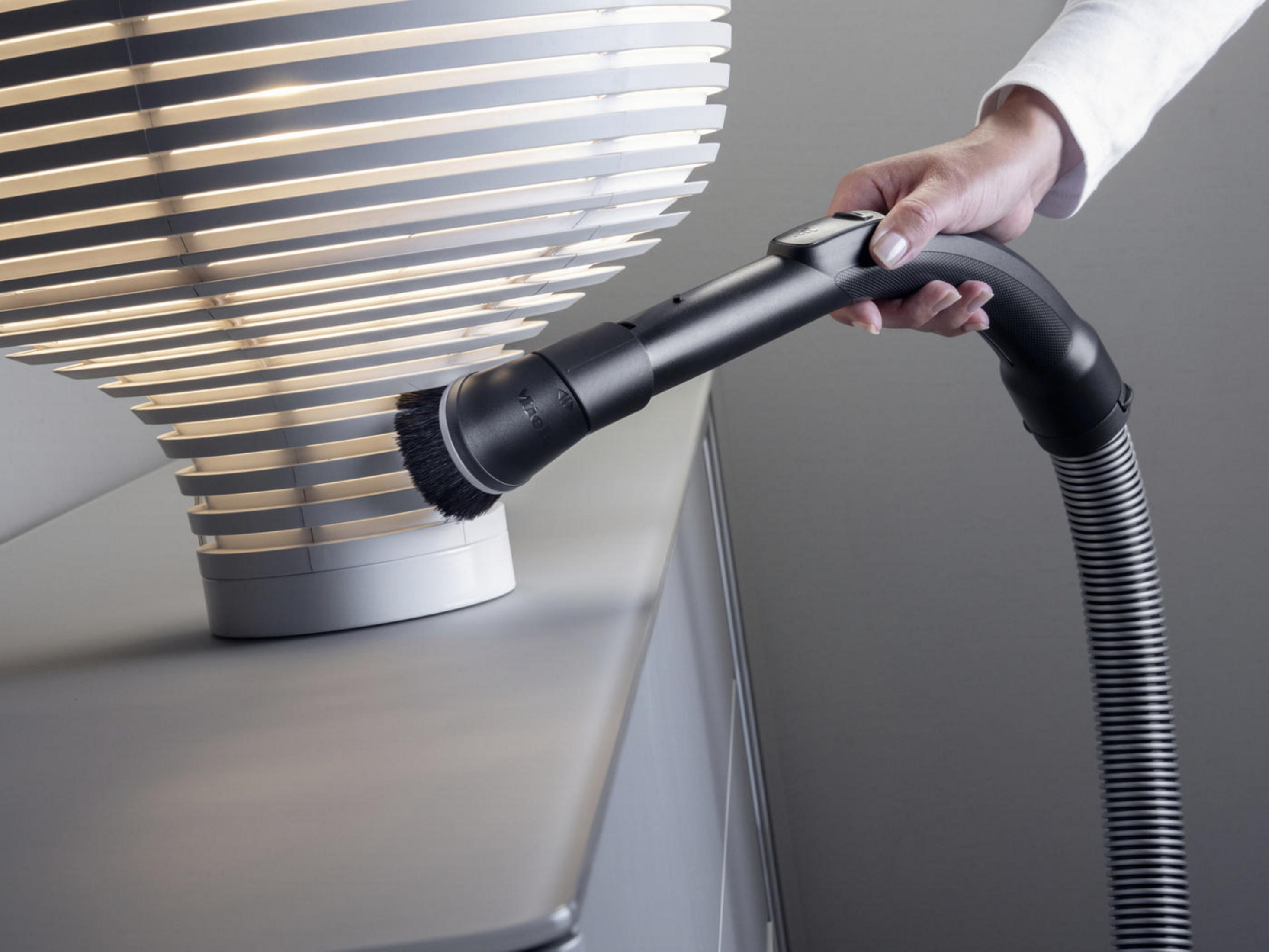 Hand benutzt Staubsaugerbürste an weißer Lampe, grauer Hintergrund.