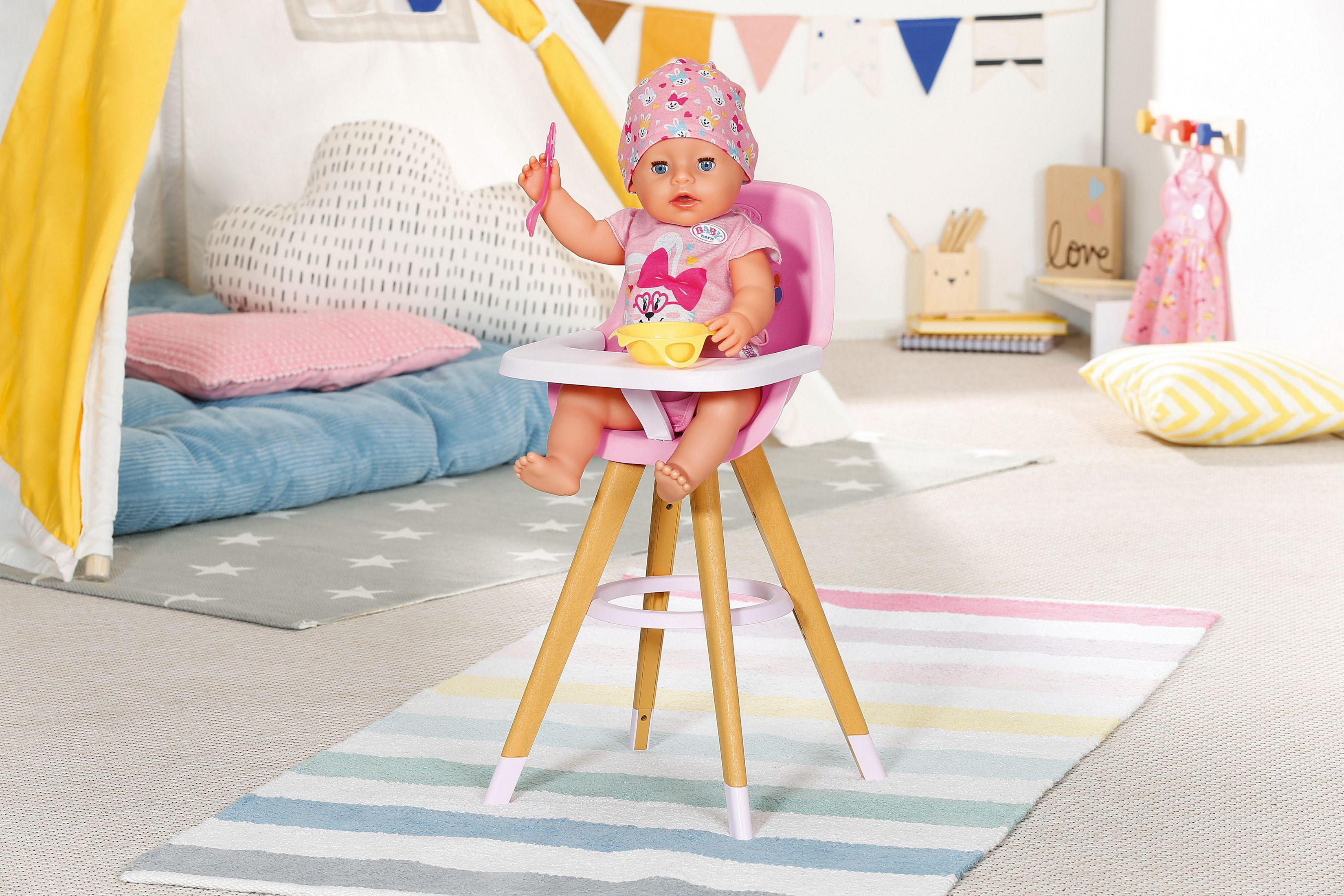 Eine Babypuppe sitzt in einem rosa Hochstuhl, hält einen Löffel, mit Spielzeug und Teppich im Bild.