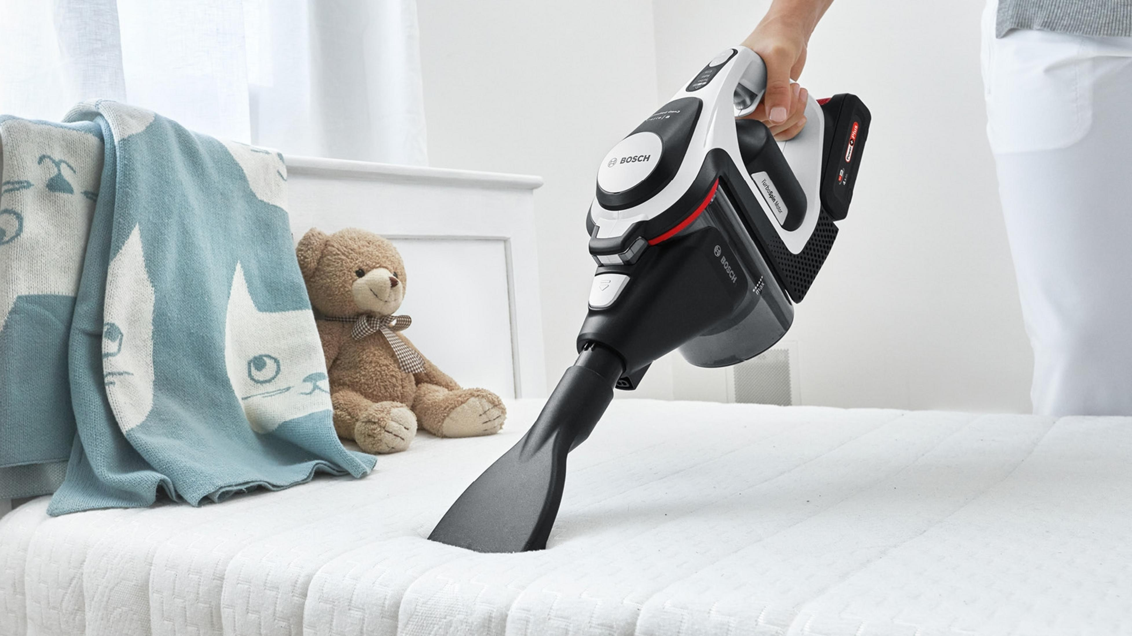 Eine Person verwendet einen kabellosen Bosch-Staubsauger auf einem Bett mit Teddybär und Decke.