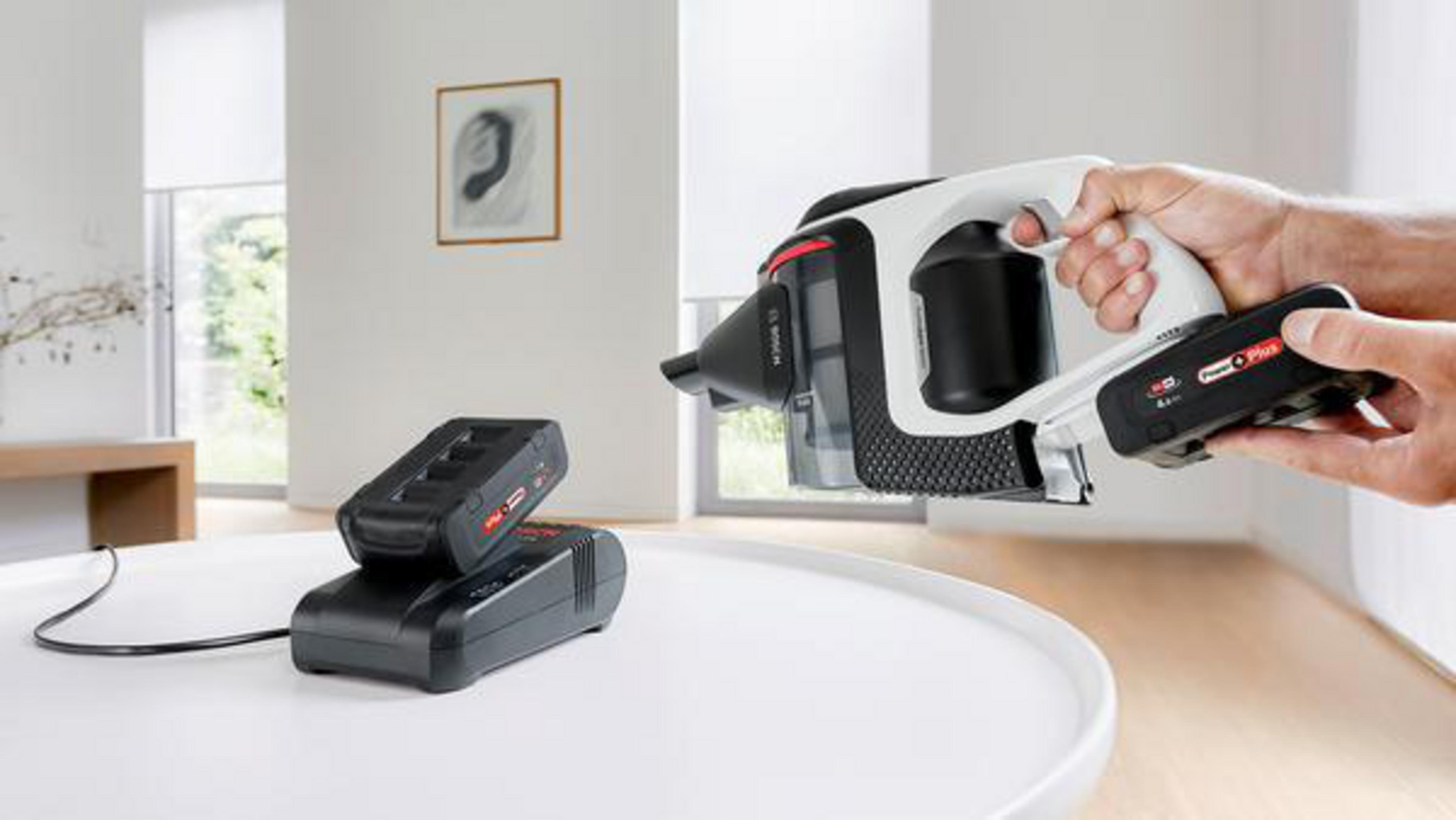 Die Hand einer Person hält einen kabellosen Staubsauger. Eine Ladestation und ein Akku sind im Vordergrund.
