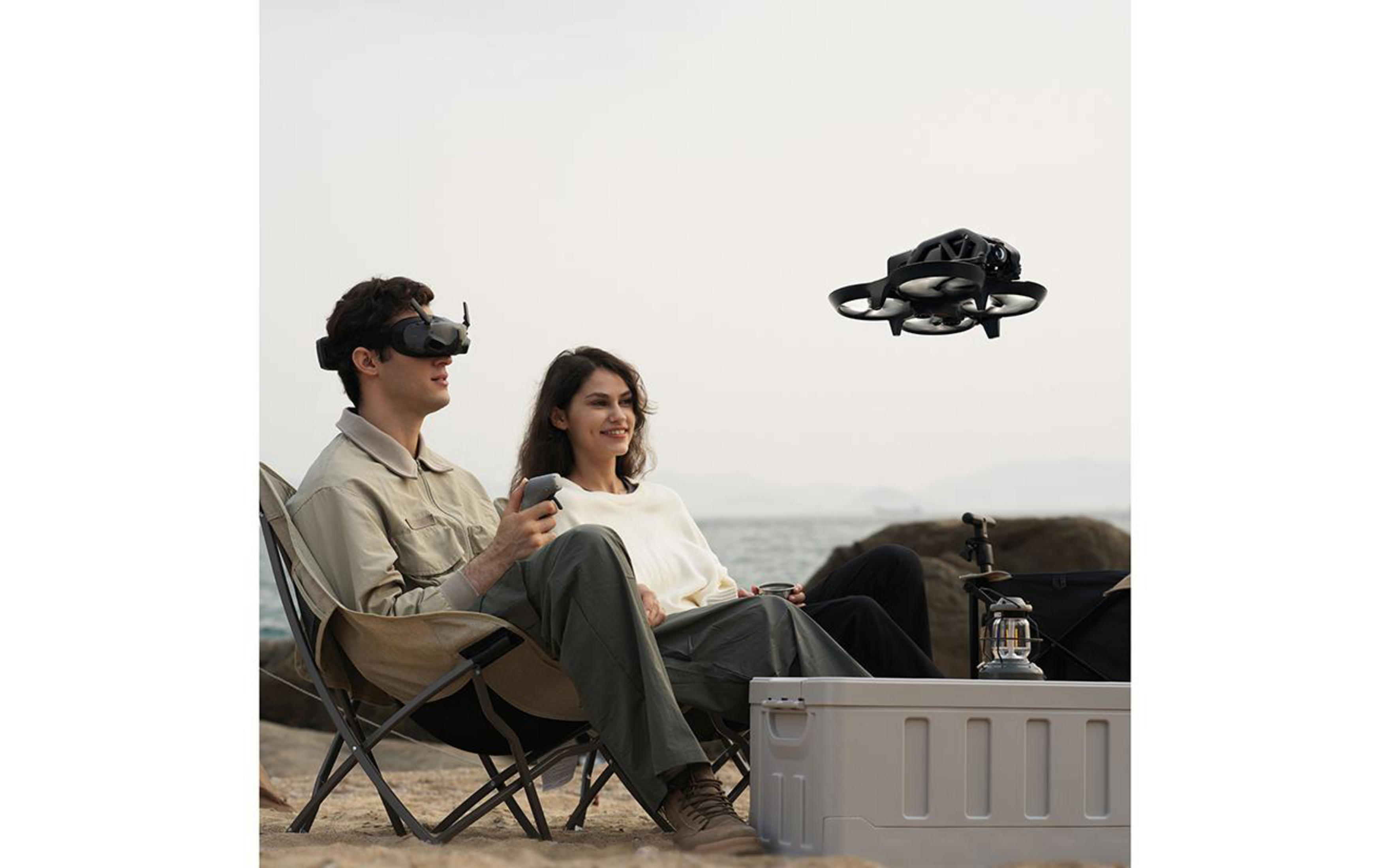 Ein Mann mit VR-Brille und eine Frau sitzen am Strand. Eine Drohne fliegt am Himmel.