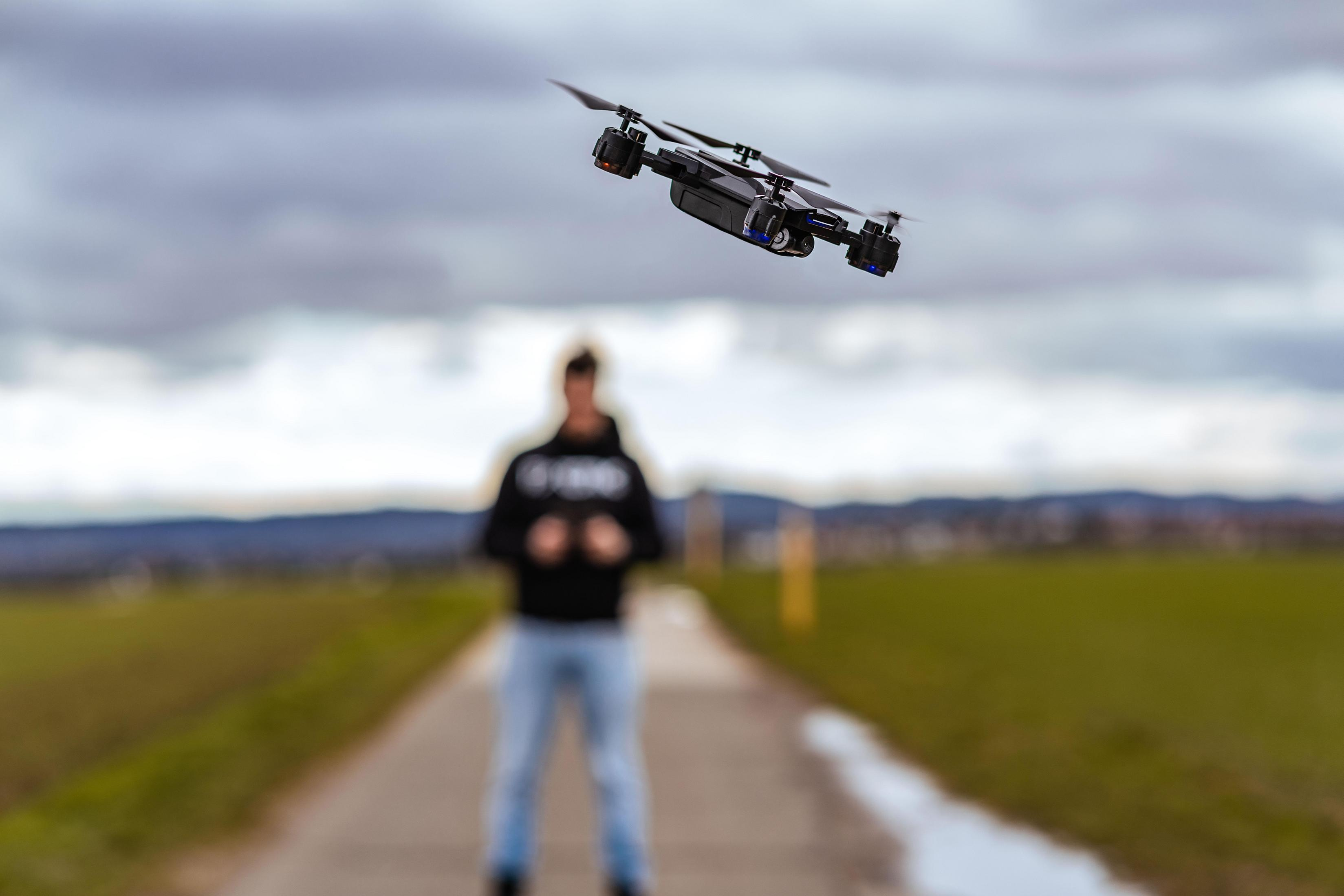 Dron w locie, z rozmytą osobą w tle. Niebo jest zachmurzone, z drogą w oddali.