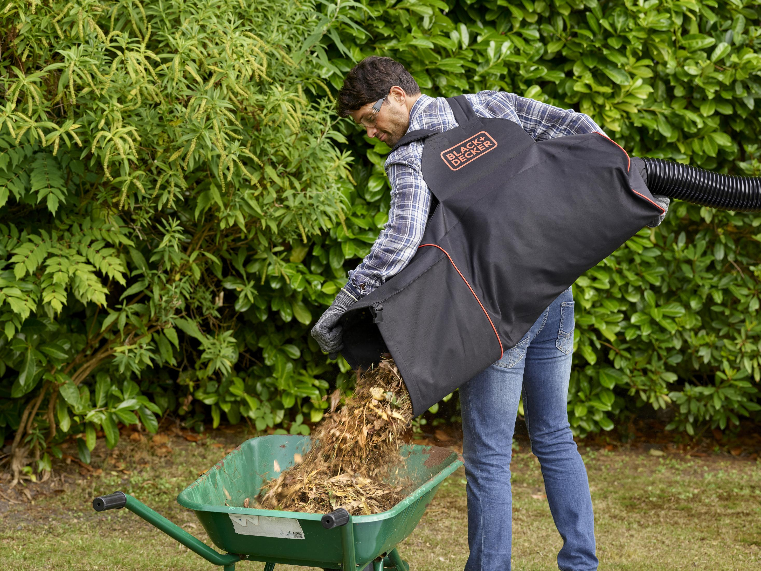 Mężczyzna opróżnia worek z liśćmi do zielonej taczki, w tle liściasty ogród.