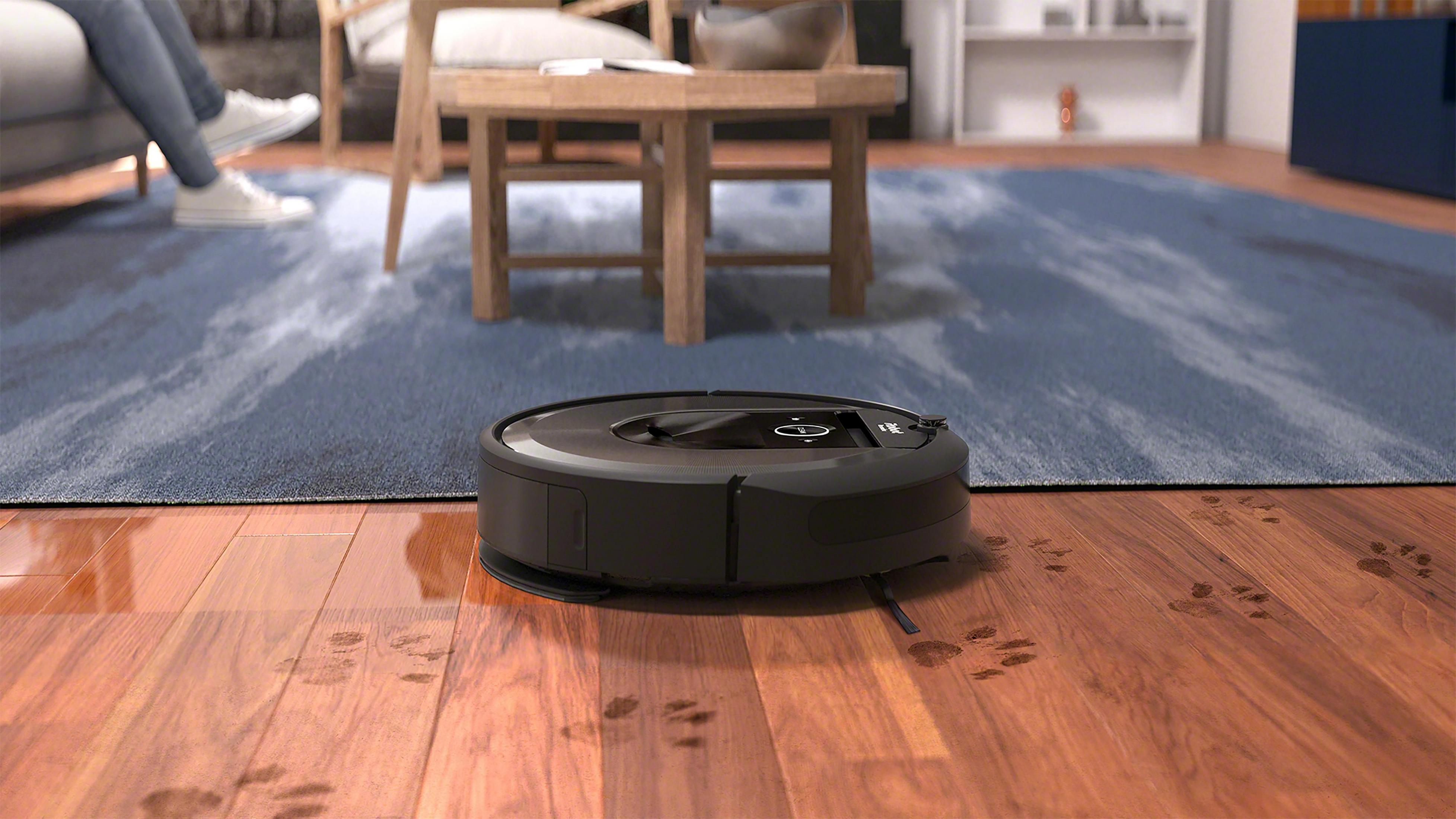 Black robot vacuum on hardwood floor. Paw prints visible. Person sitting in background.