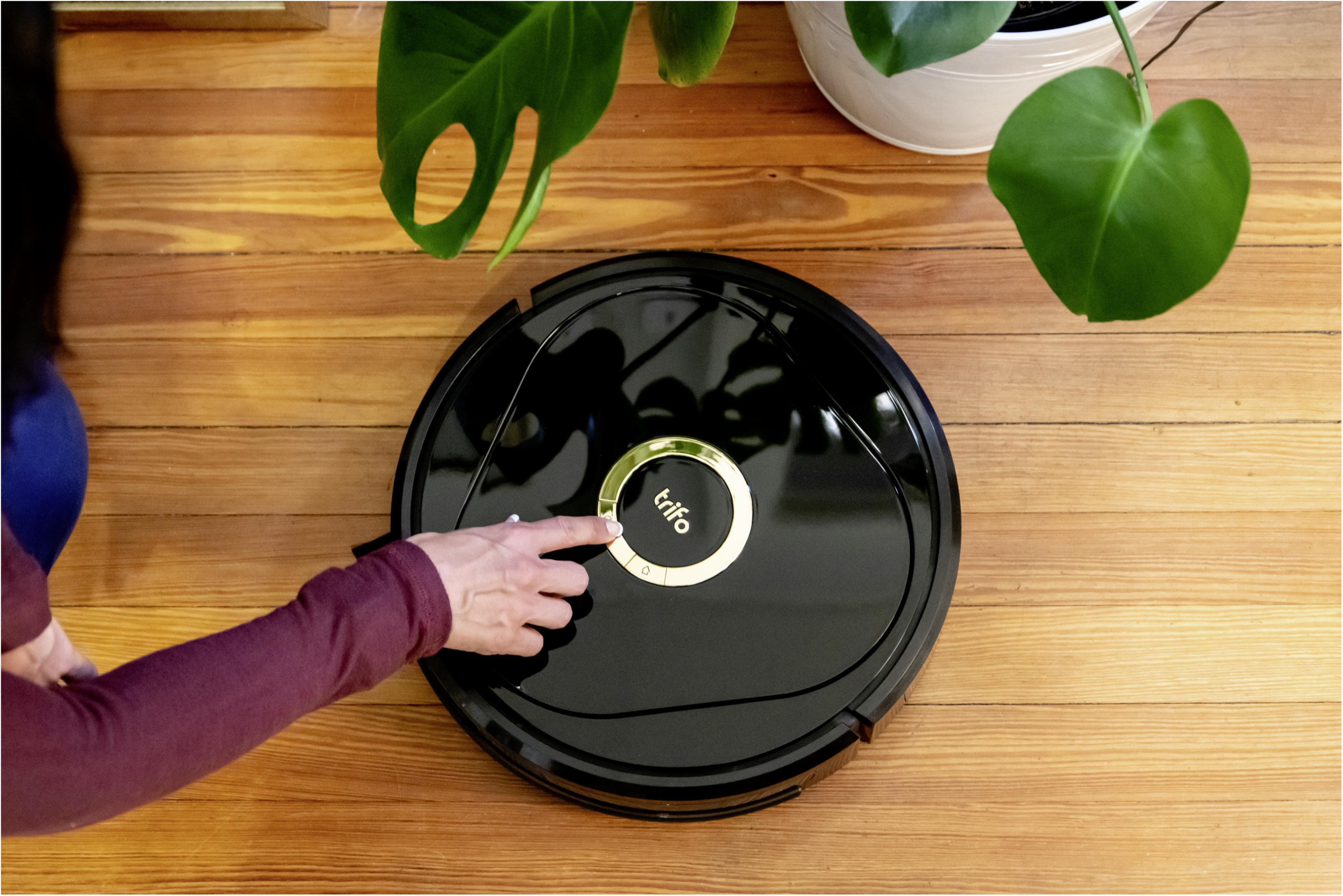 Ręka osoby naciska przycisk na czarnym robocie odkurzaczu. Roślina doniczkowa w tle.