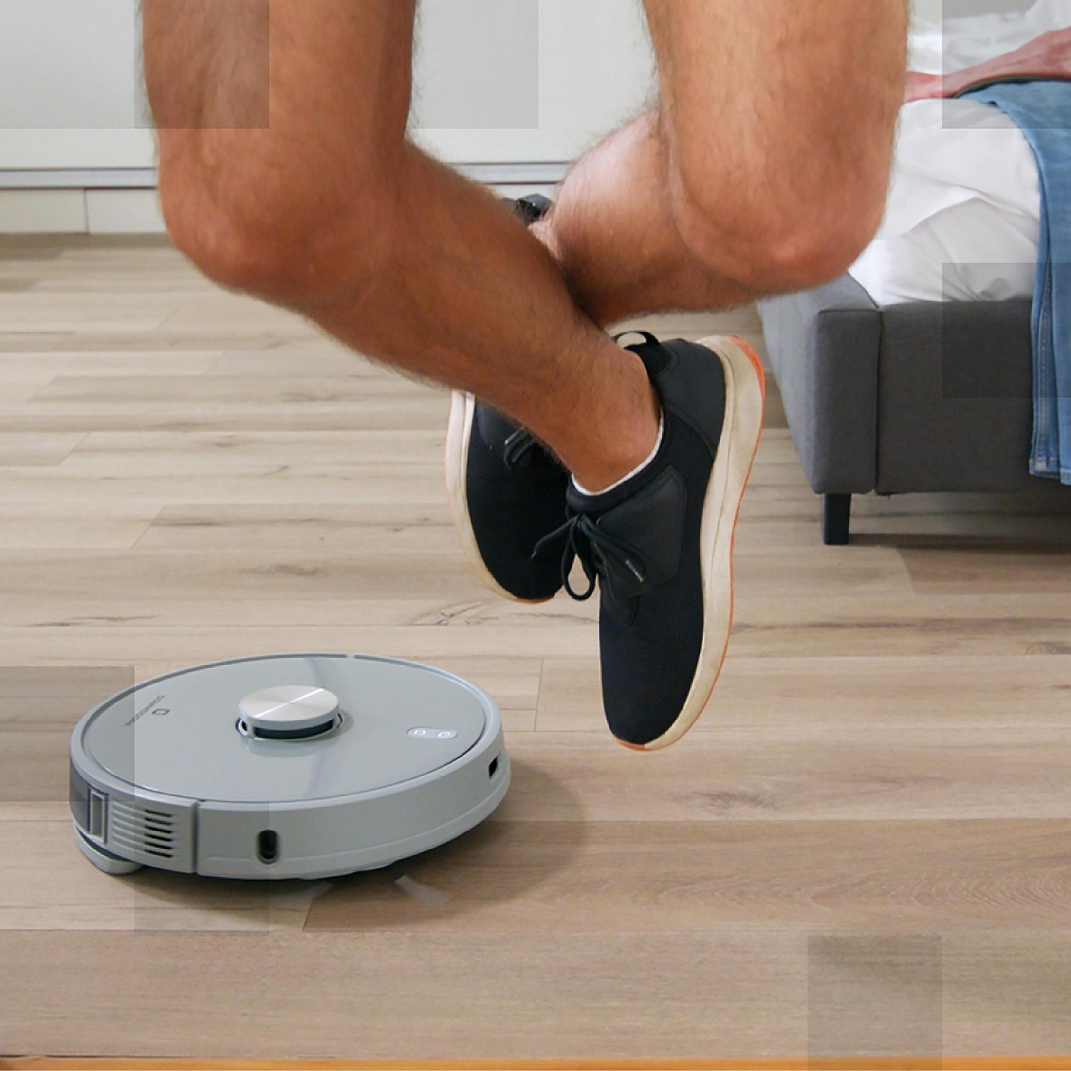 Person springt über Roboter-Staubsauger auf Holzboden.