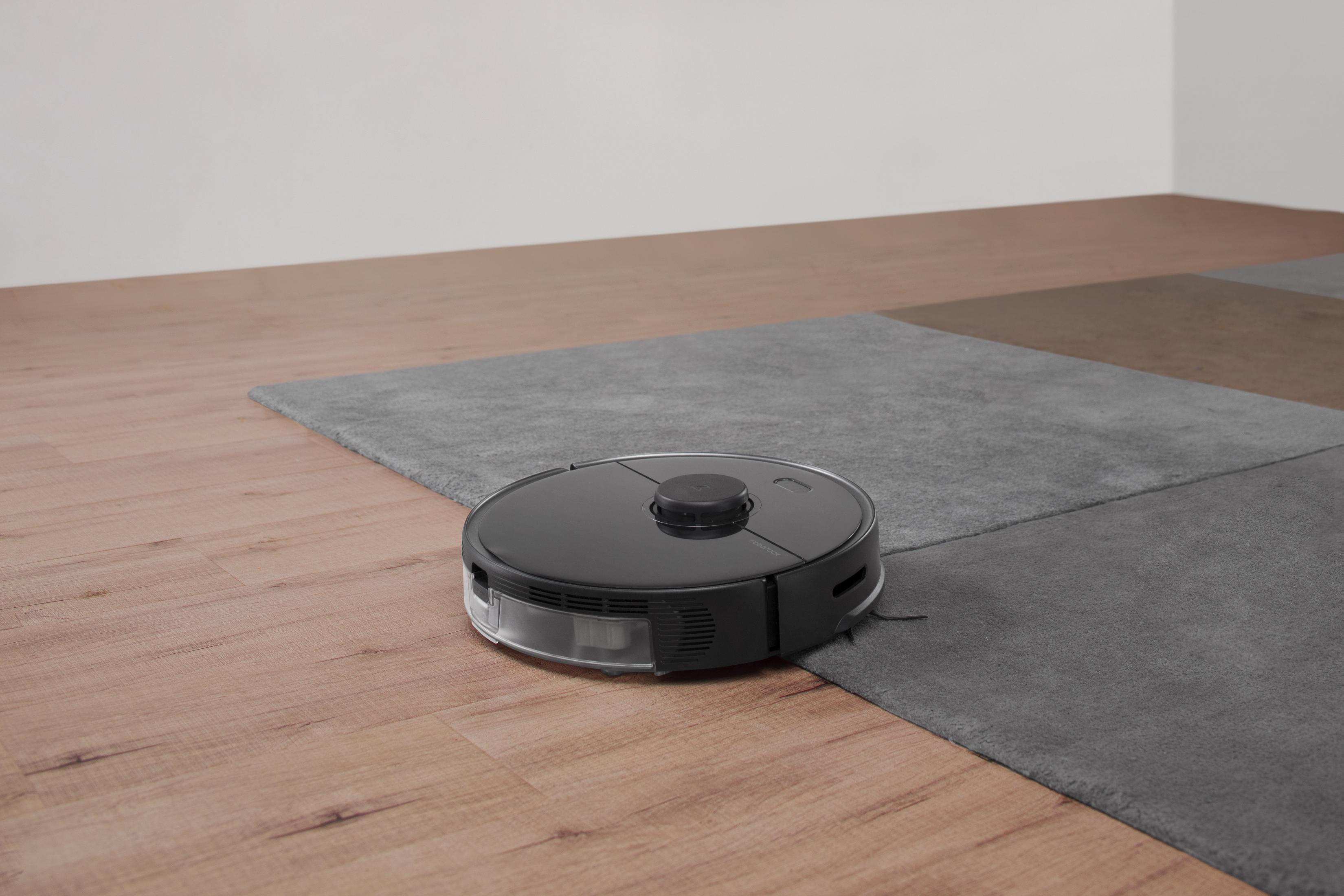 Ein schwarzer Roboter-Staubsauger auf einem Holzboden, der auf einen grauen Teppich übergeht.