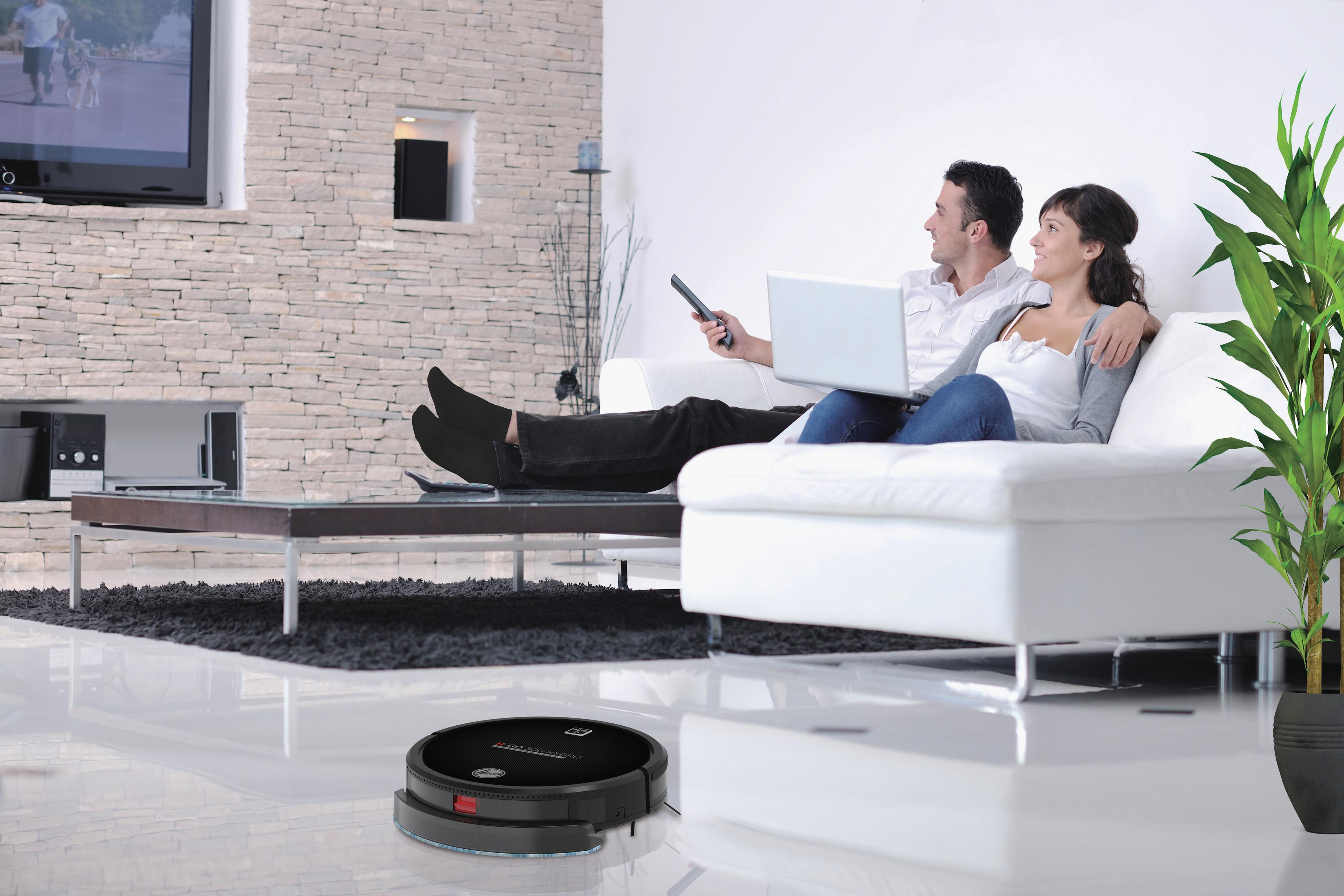 Ein Paar sitzt auf einer Couch und schaut fern. Ein Roboter-Staubsauger liegt auf dem Boden.