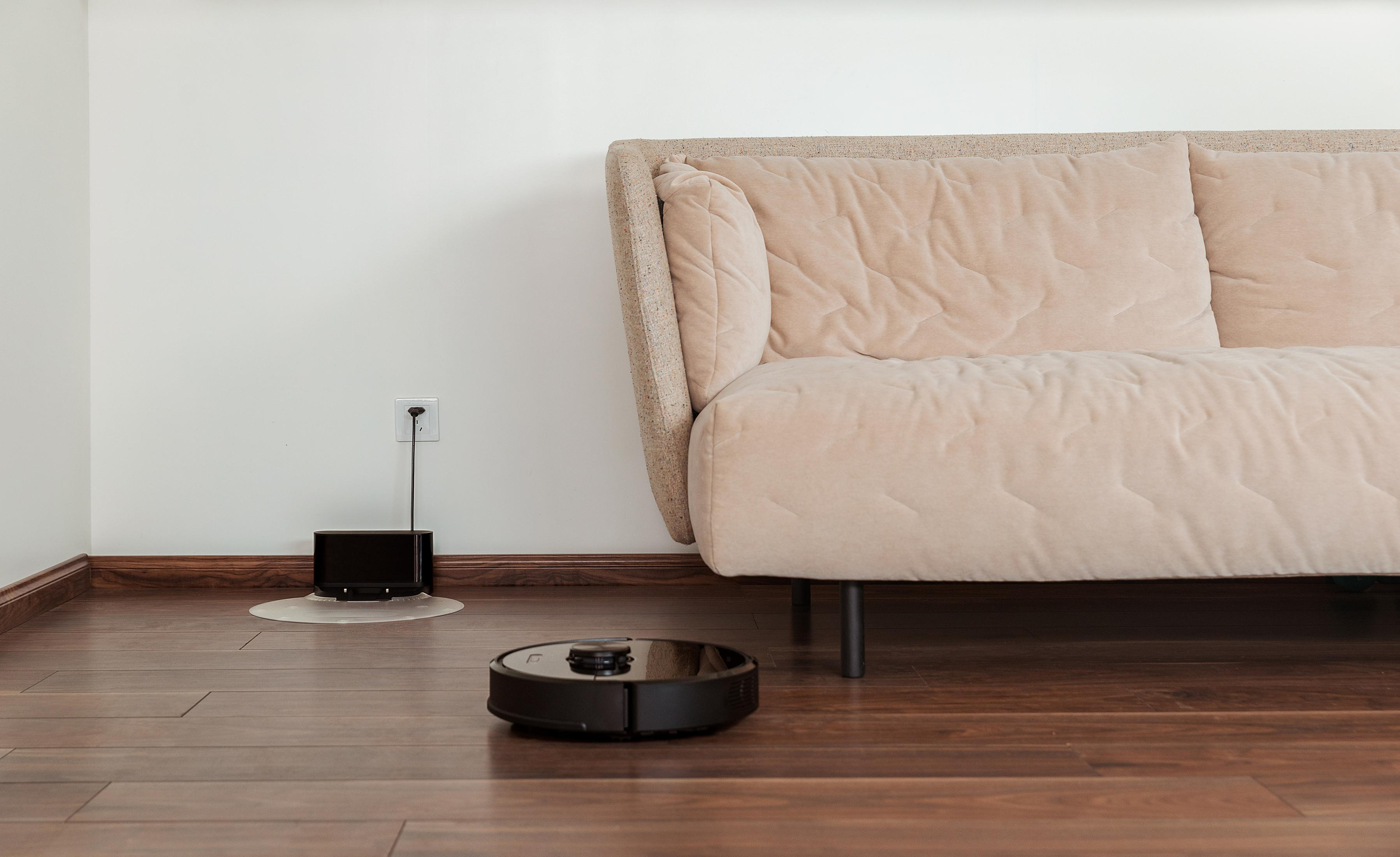 Schwarzer Roboter-Staubsauger auf Holzboden. Beige Sofa und weiße Wand im Hintergrund. Steckdose.