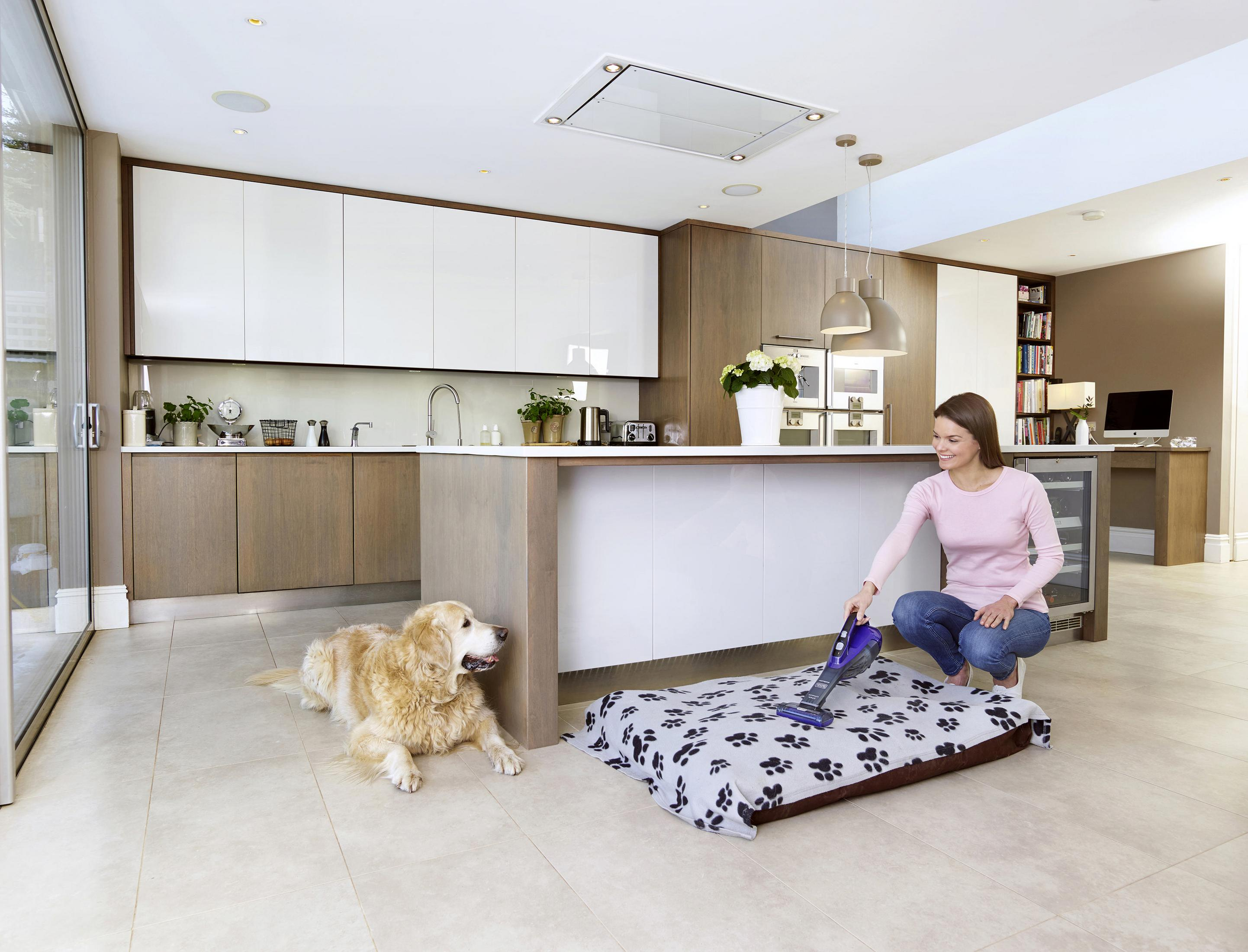 Mujer aspirando una cama para perros con un perro cerca. Fondo de cocina moderna.