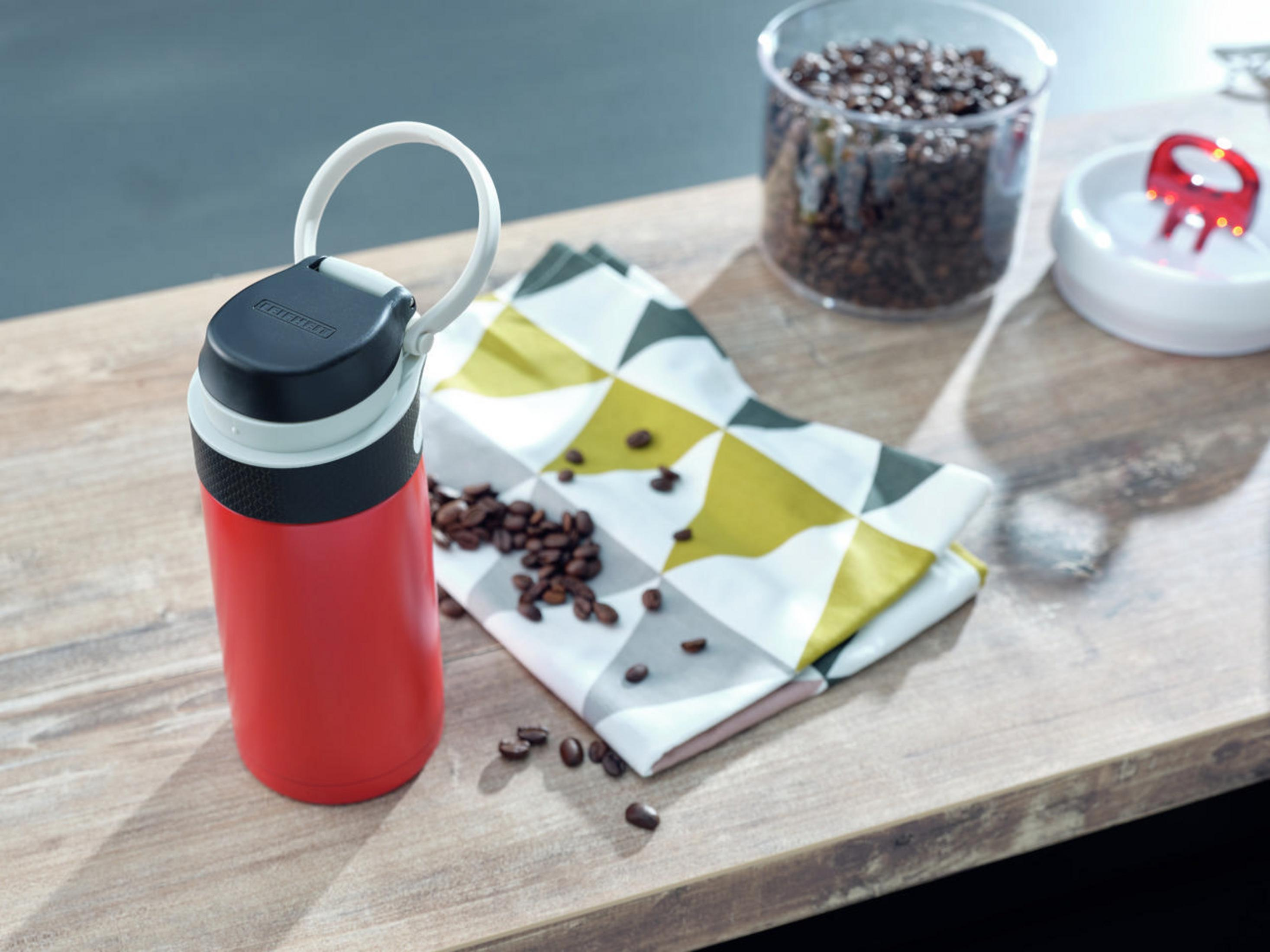 Rote und schwarze Isolierkanne neben Kaffeebohnen, einer Serviette und einem Glas.