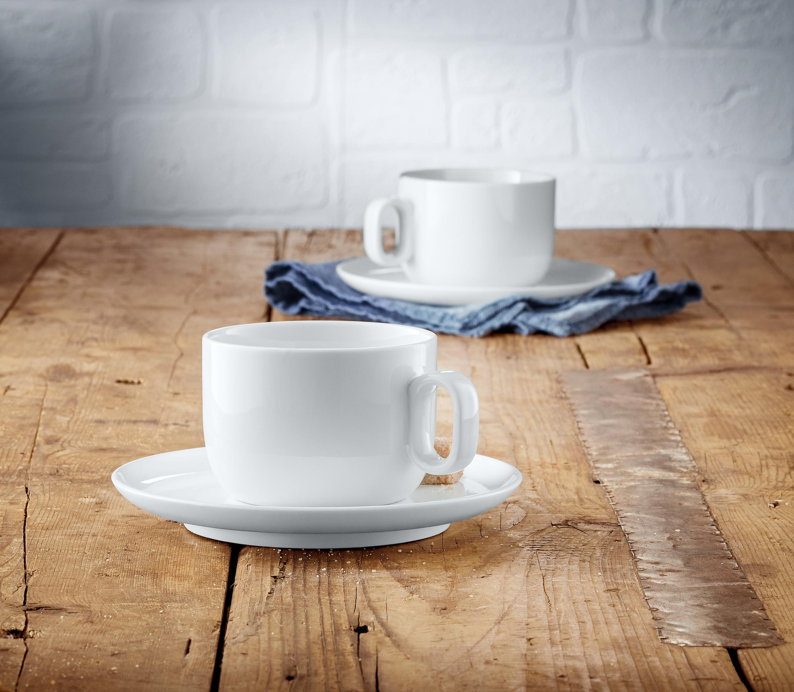 Zwei weiße Kaffeetassen und Untertassen auf einem Holztisch. Im Hintergrund liegt eine blaue Serviette.
