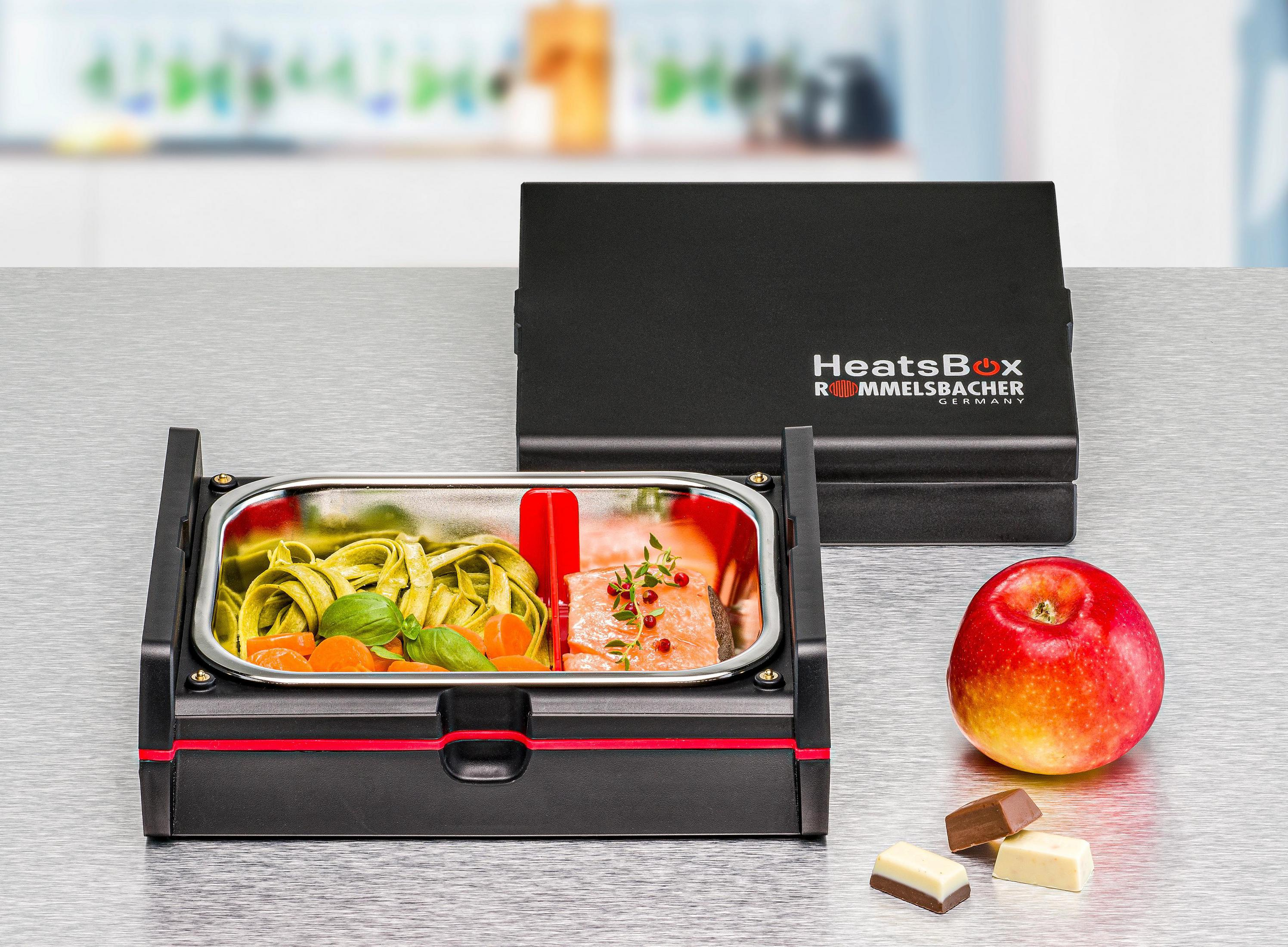 Eine schwarze beheizbare Lunchbox mit Essen und geschlossenem Deckel. Apfel und Schokolade daneben.