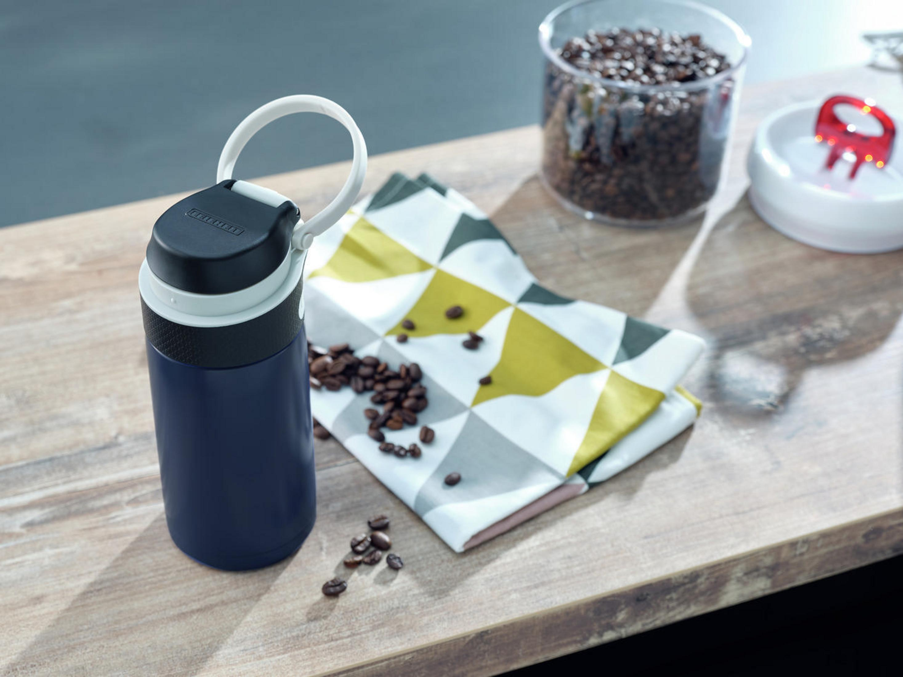 Eine blau-schwarze Thermoskanne mit weißem Deckel und Griff. Kaffeebohnen und ein Handtuch sind in der Nähe.