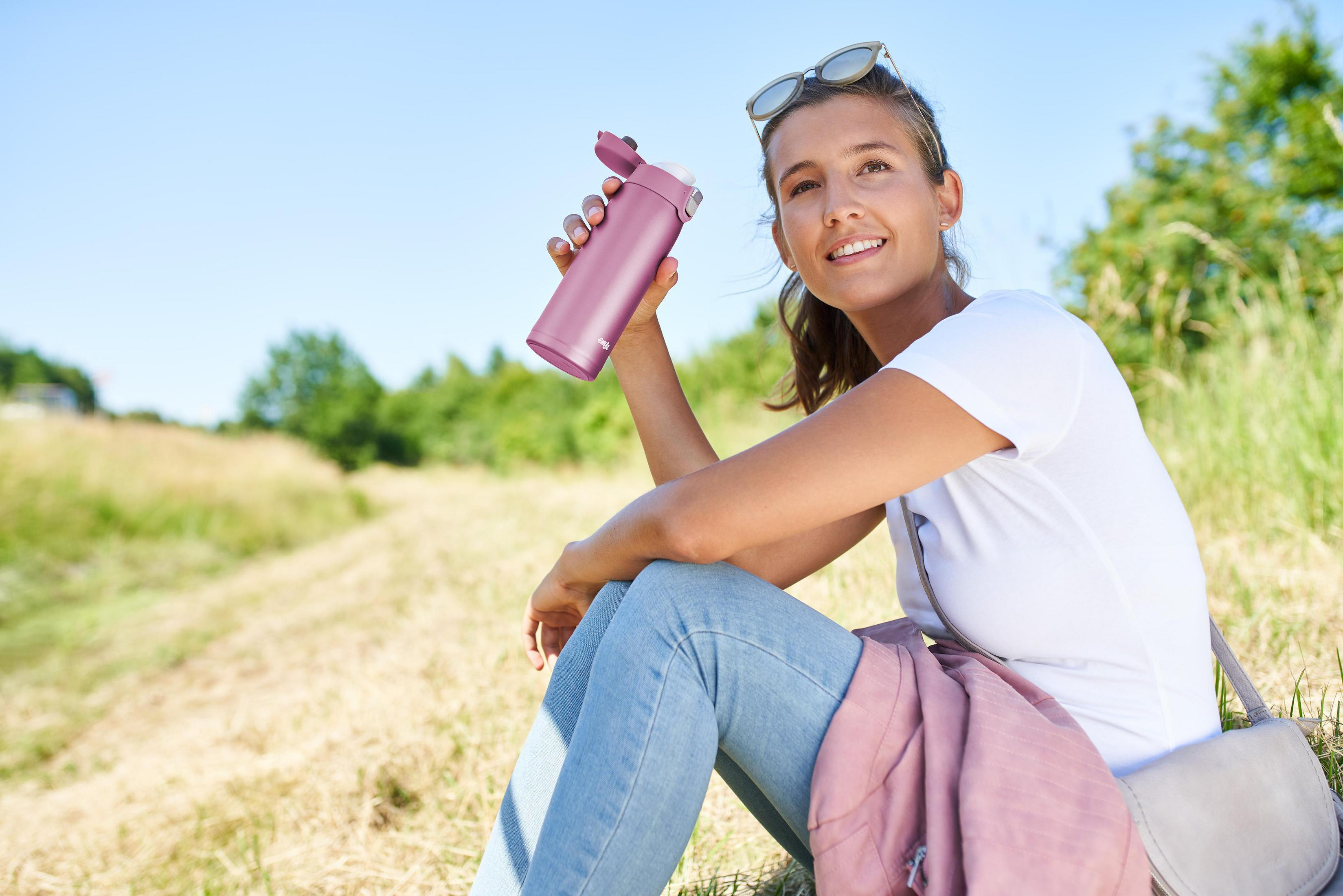 Frau mit rosa Wasserflasche, sitzt im Gras an einem sonnigen Tag.