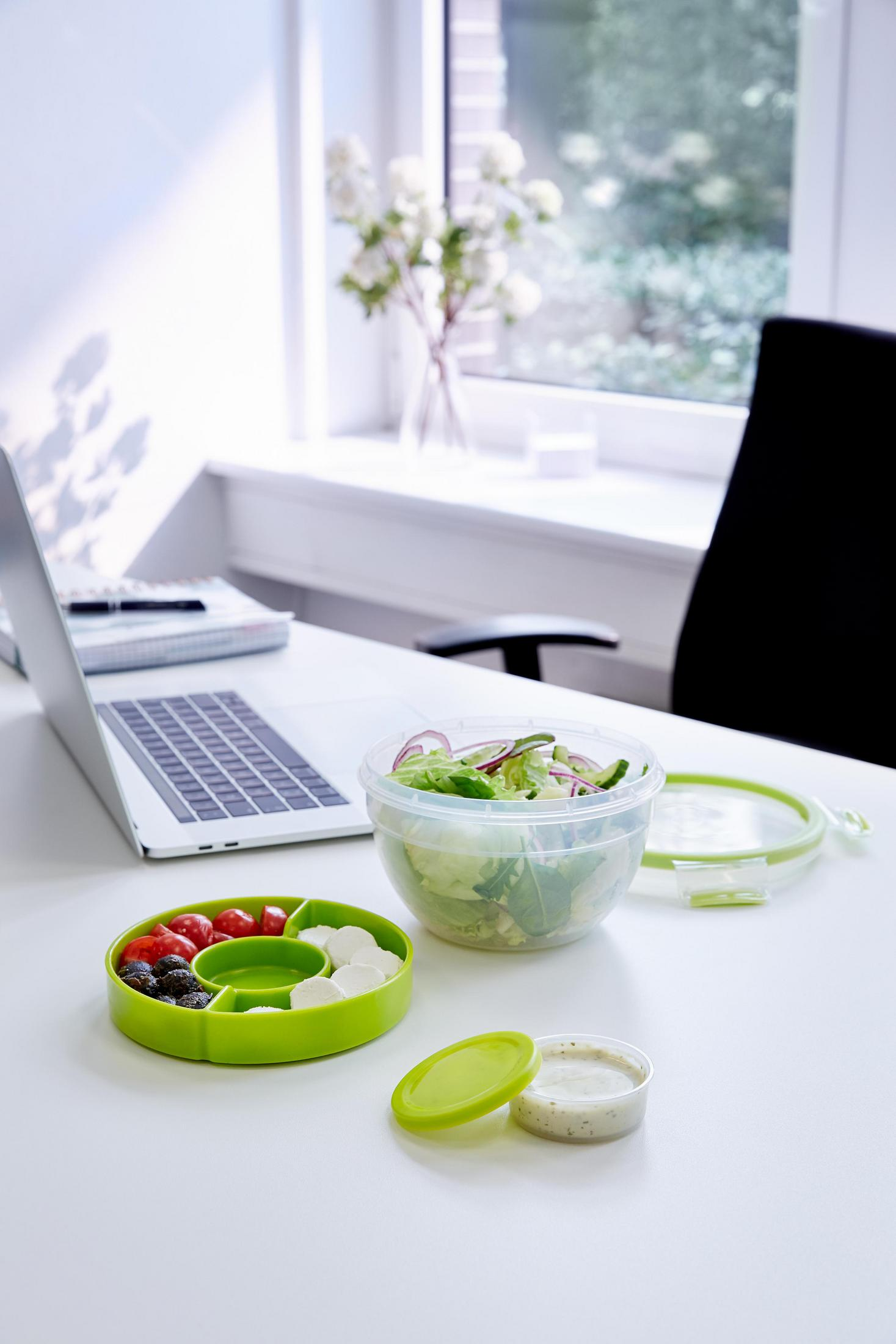 Salatbehälter mit Salat, Tomaten und Mozzarella-Kugeln. Laptop im Hintergrund. Weißer Tisch.