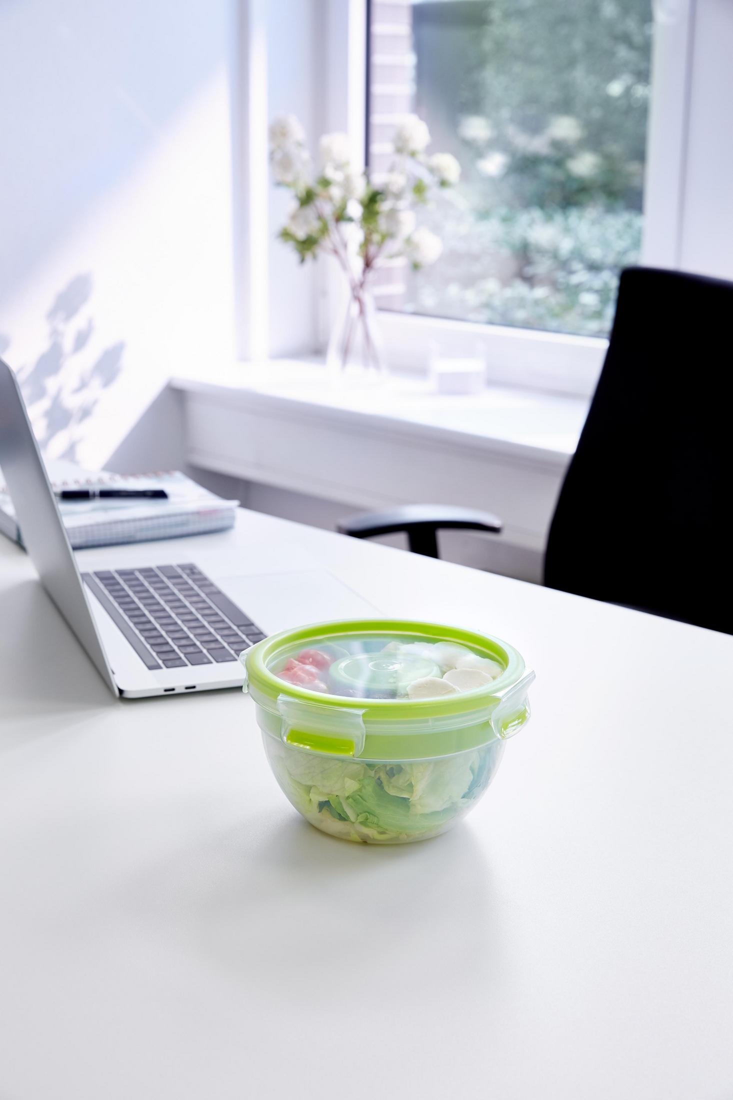 Ein Salat in einem transparenten Behälter auf einem weißen Schreibtisch. Ein Laptop und Blumen sind im Hintergrund.
