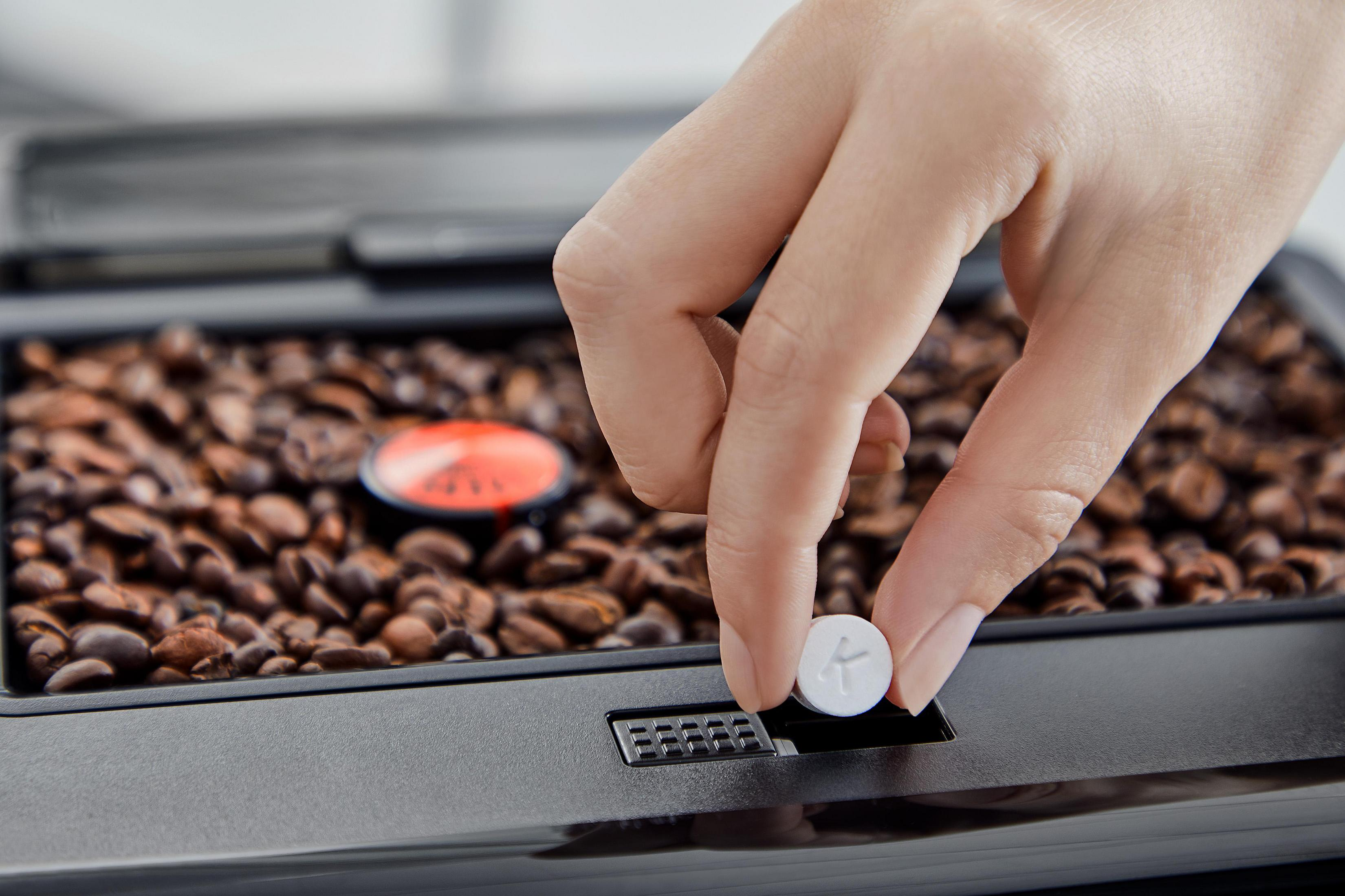 Ręka wkłada białą tabletkę do ekspresu do kawy z ziarnami kawy.