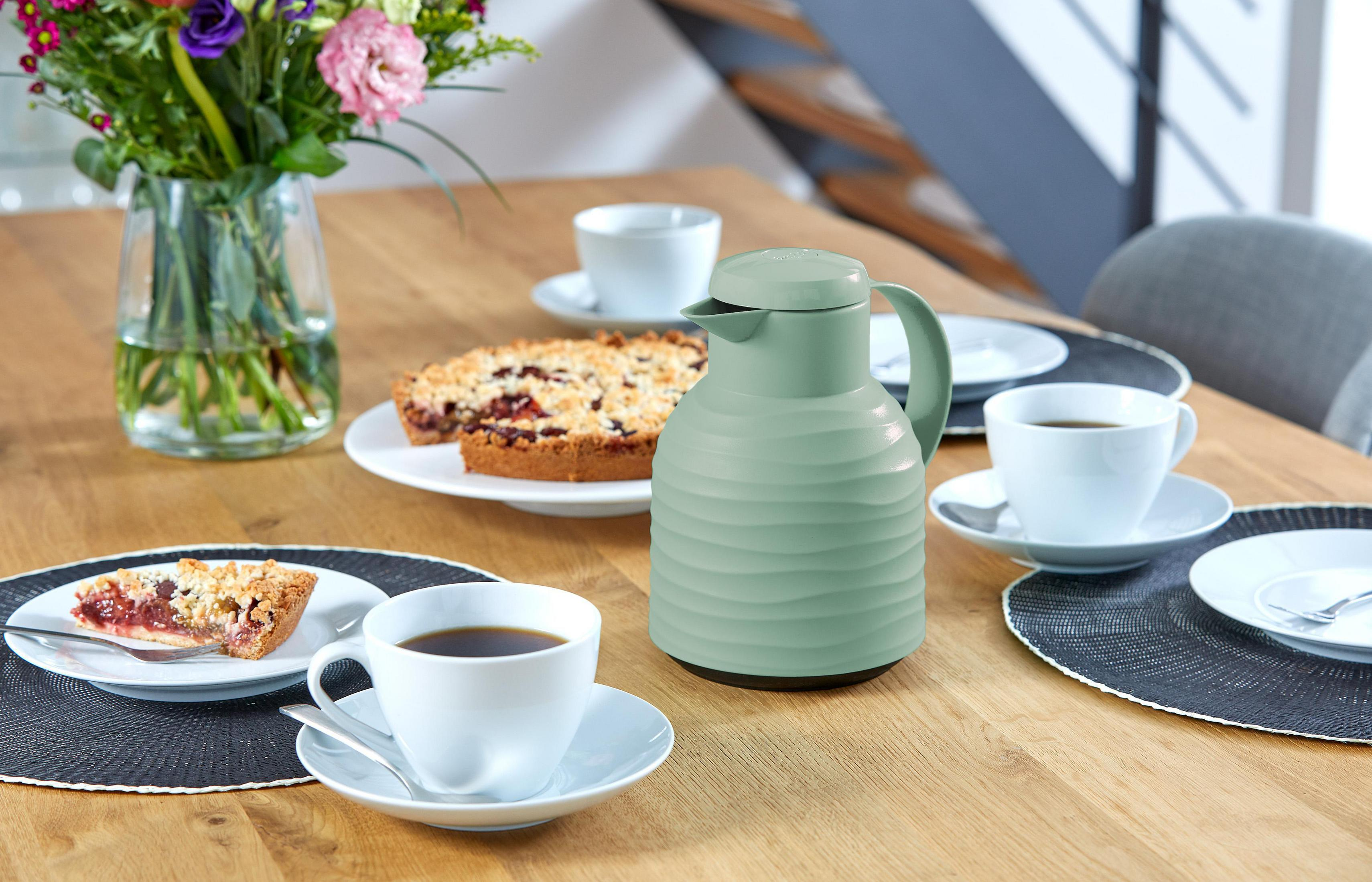 Tischgedeck. Eine grüne Thermoskanne, Kaffeetassen und Gebäck stehen auf dem Holztisch. Blumen sind in einer Vase.