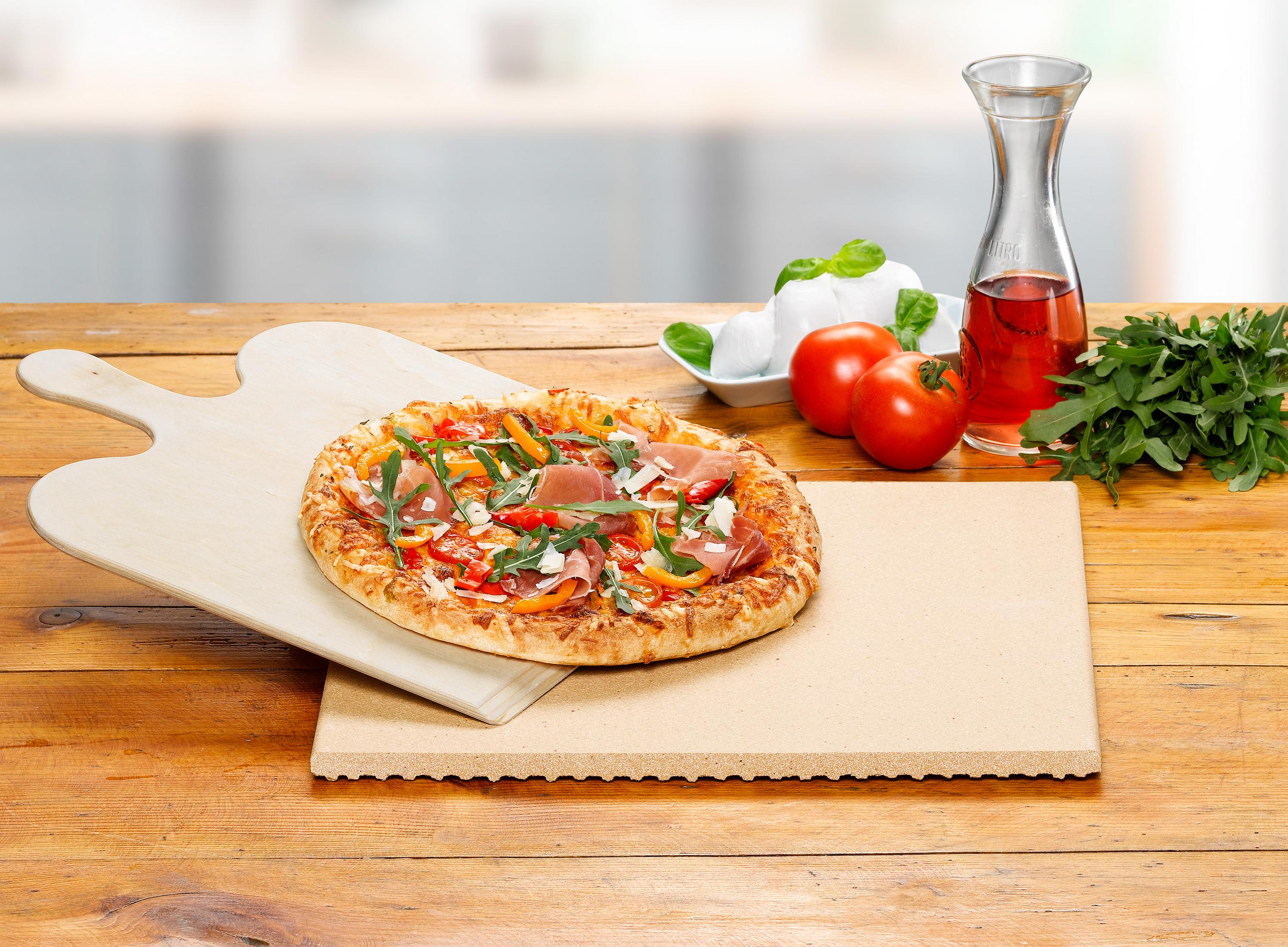 Eine Pizza auf einem Holzpaddel mit Zutaten, neben einem Steinbackbrett, auf einem Tisch.