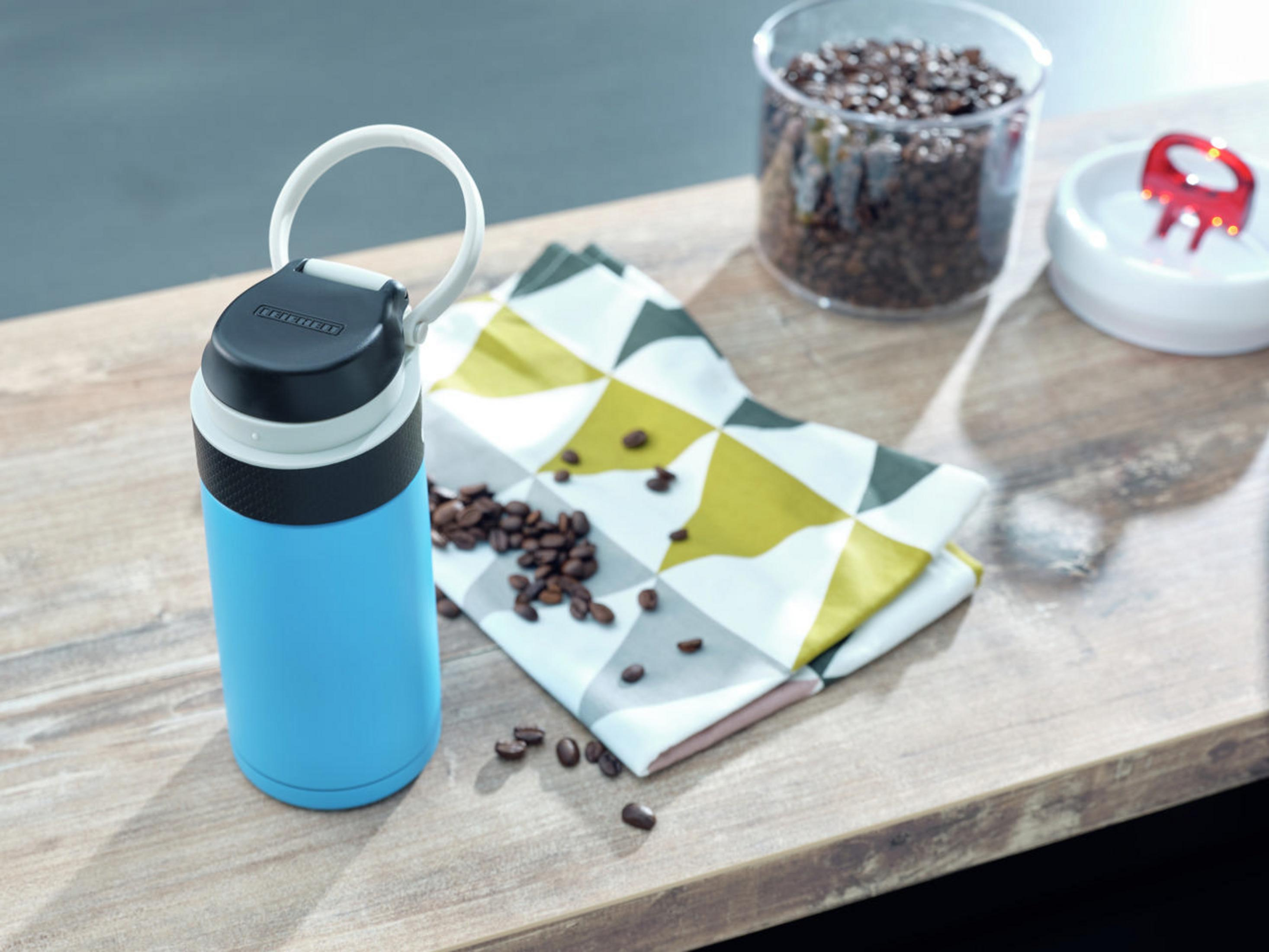 Eine blaue Thermosflasche, Kaffeebohnen auf einer Serviette und ein durchsichtiges Glas mit Bohnen auf Holz.
