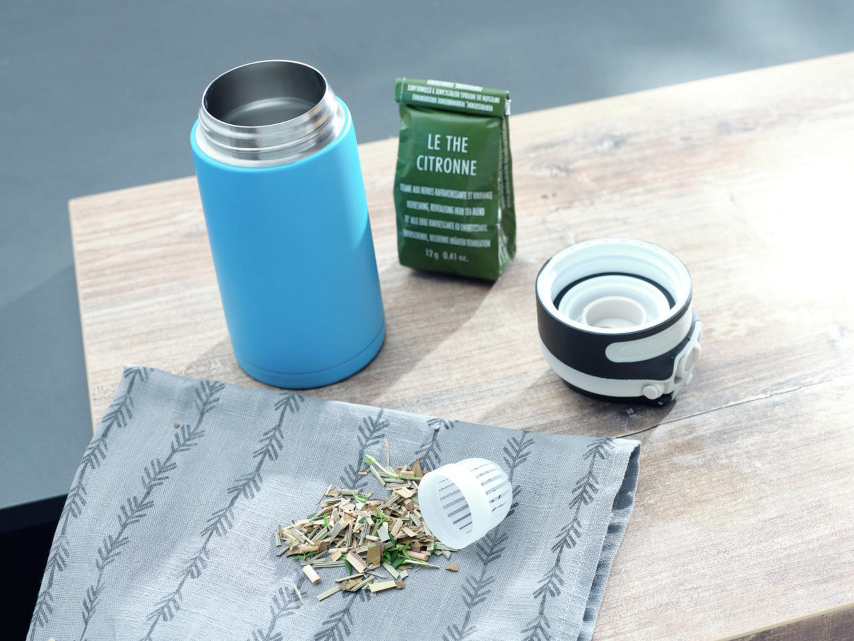 Eine blaue Isolierflasche, ein grüner Teebeutel, ein Tee-Aufgussgerät und Teeblätter auf einem Holztisch.