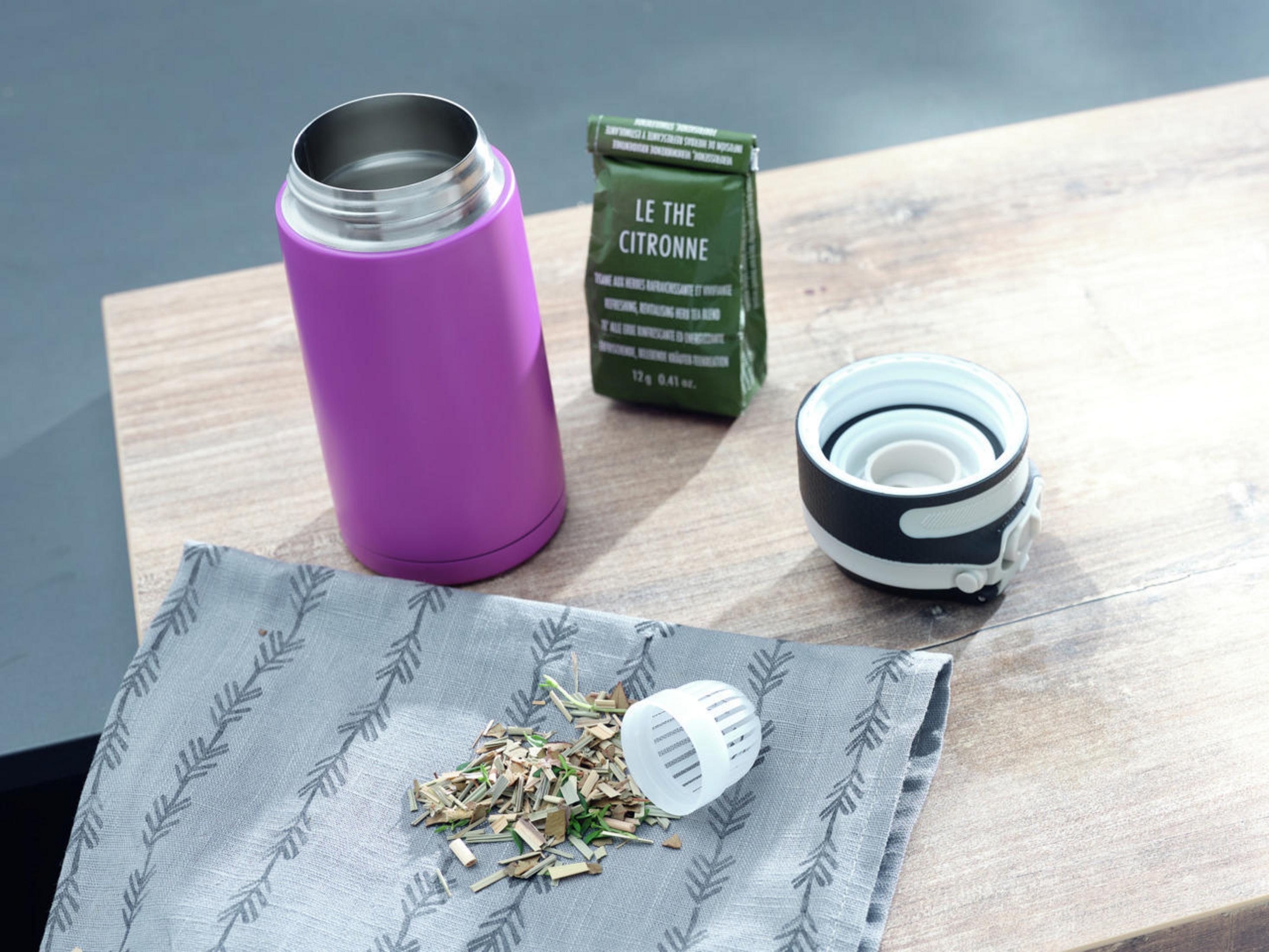 Violette Isolierflasche, Teeblätter, Tee-Einsatz und Teebeutel auf Holzoberfläche.