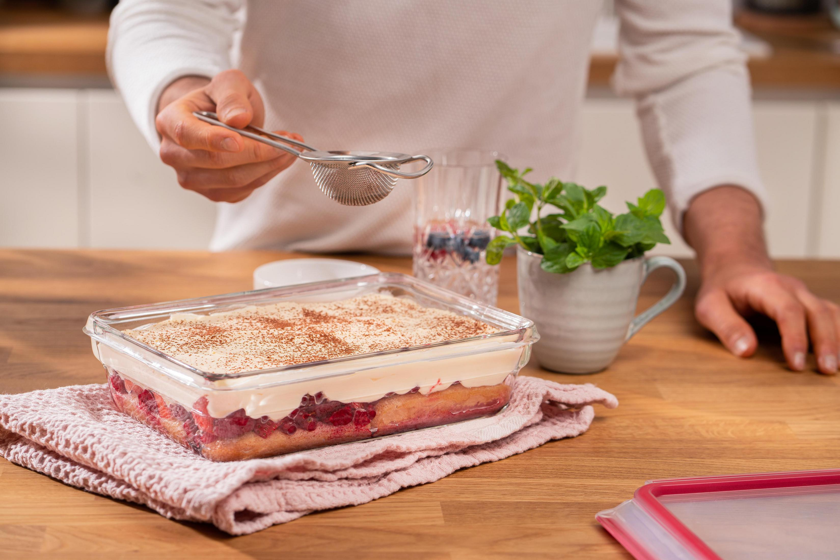 Eine Person streut Puderzucker auf ein Dessert in einer Glasschale. Minze und Beeren liegen daneben.