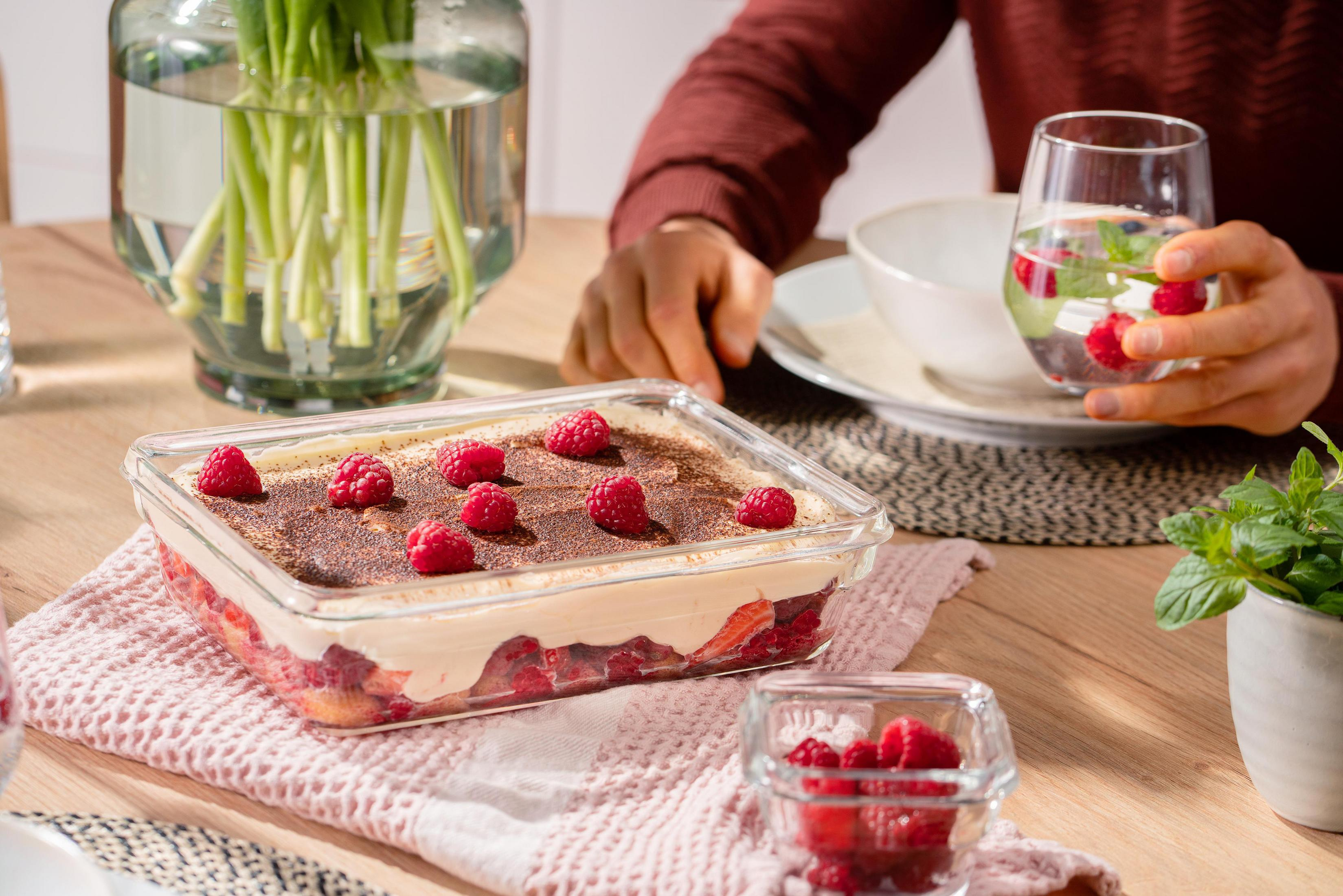 Himbeer-Tiramisu in einer Glasschale, Person hält ein Wasserglas. Anderes Essen und Blumen sind auf dem Tisch.