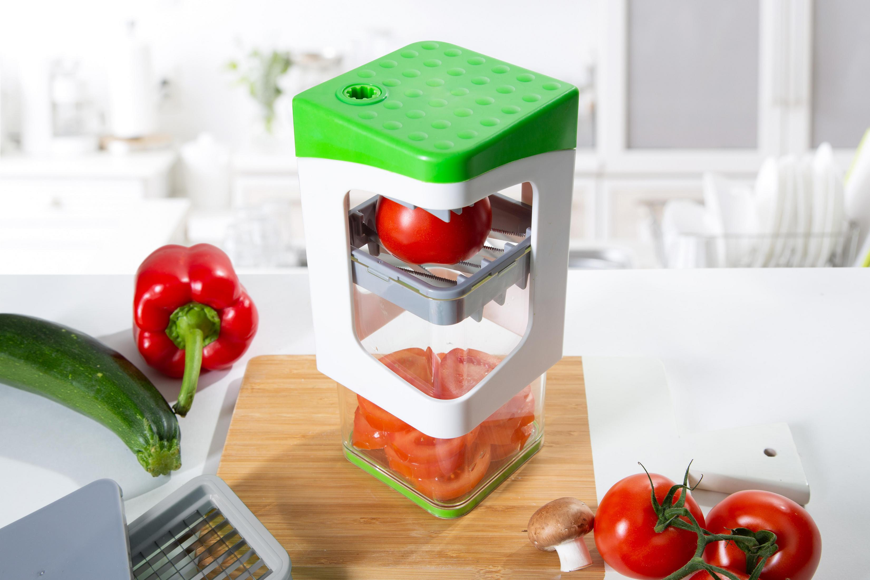 Eine Tomate wird mit einem grün-weißen Küchenwerkzeug geschnitten, andere Gemüse im Bild.