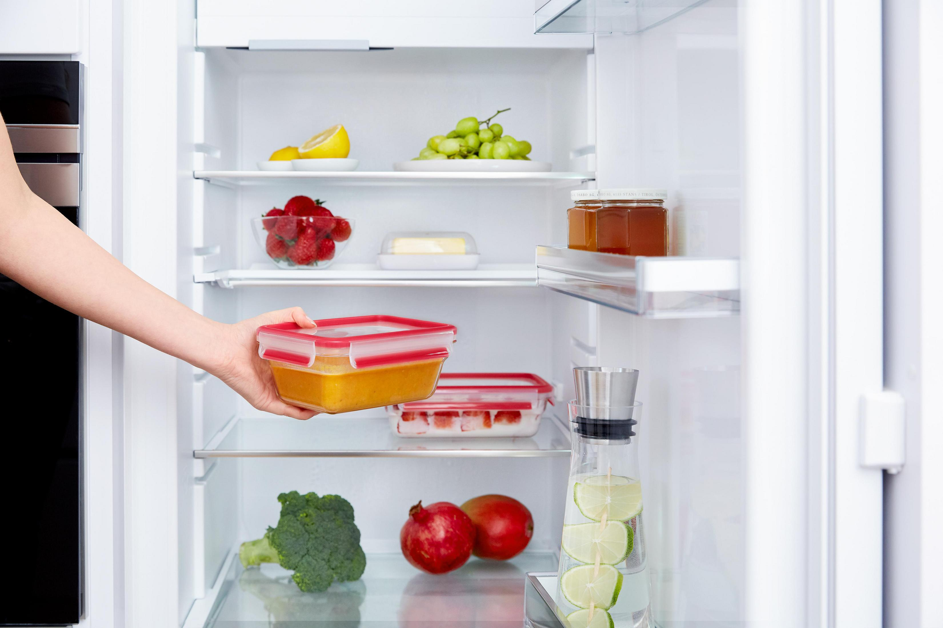 Hand legt einen Behälter mit Essen in einen Kühlschrank mit Obst und anderen Lebensmitteln.
