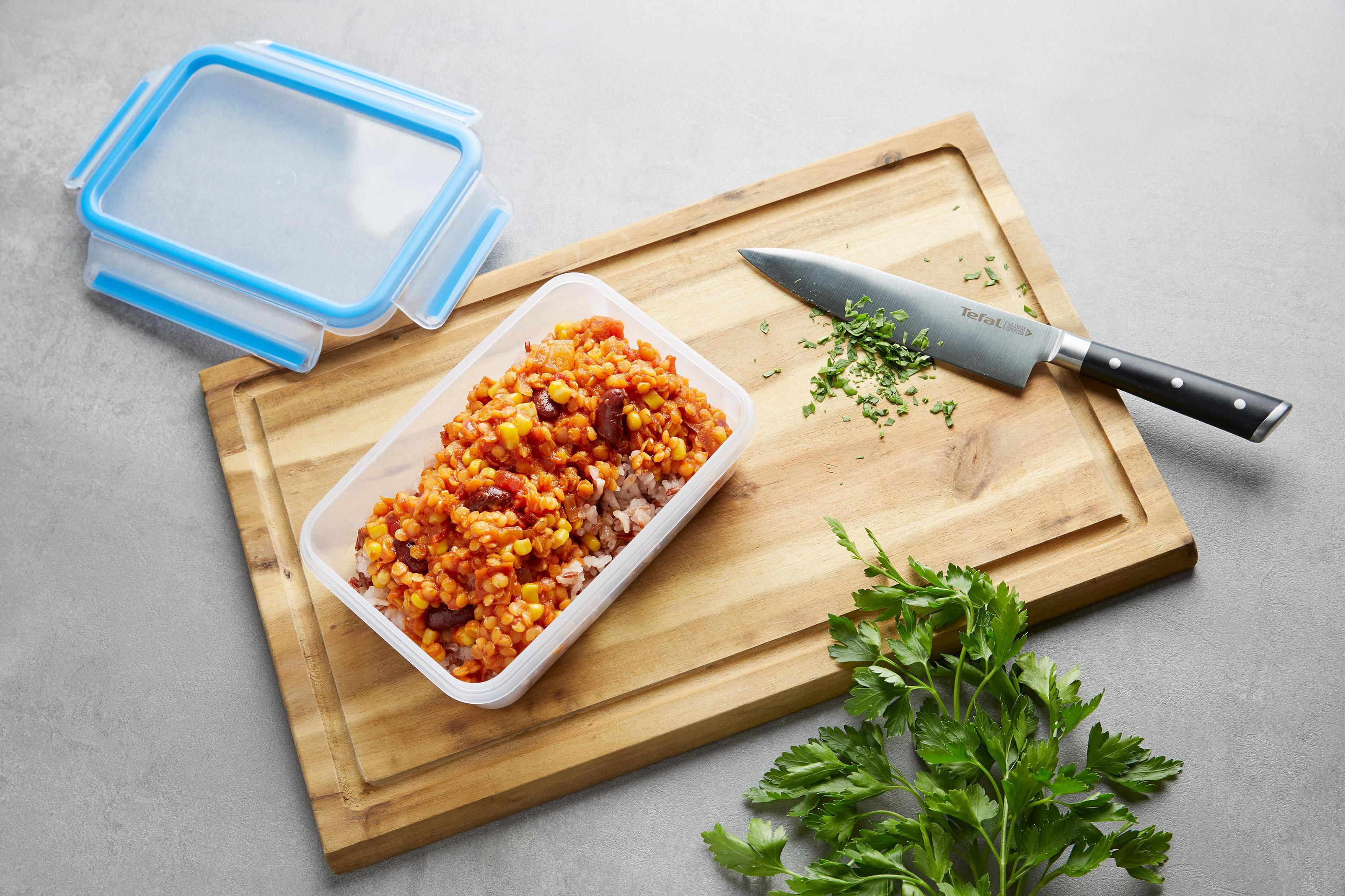 Mahlzeit in einem Plastikbehälter mit Messer und Petersilie auf einem Holzbrett. Grauer Hintergrund.