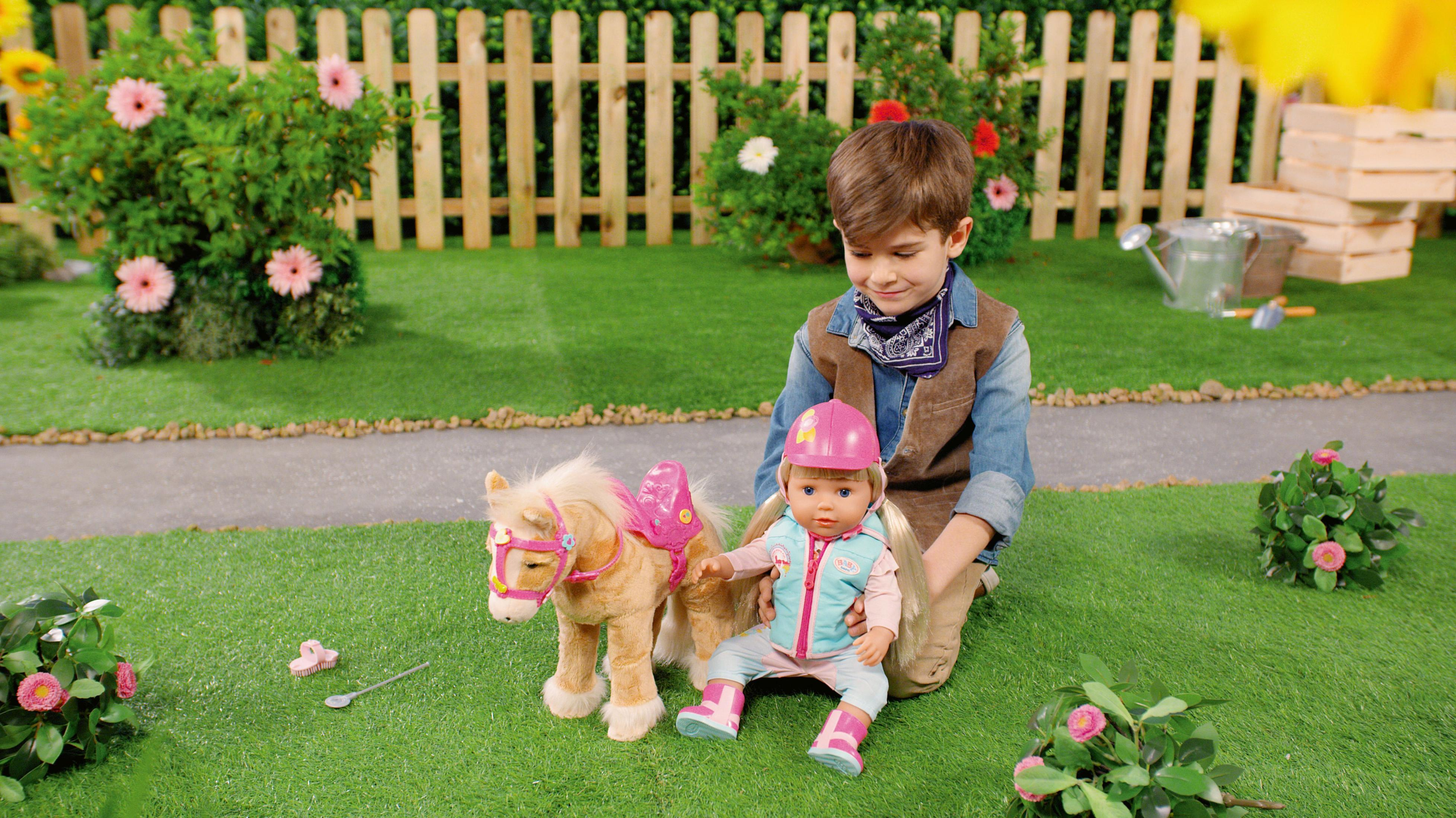 Junge mit Puppe und Spielzeugpferd auf Gras, Zaun im Hintergrund.