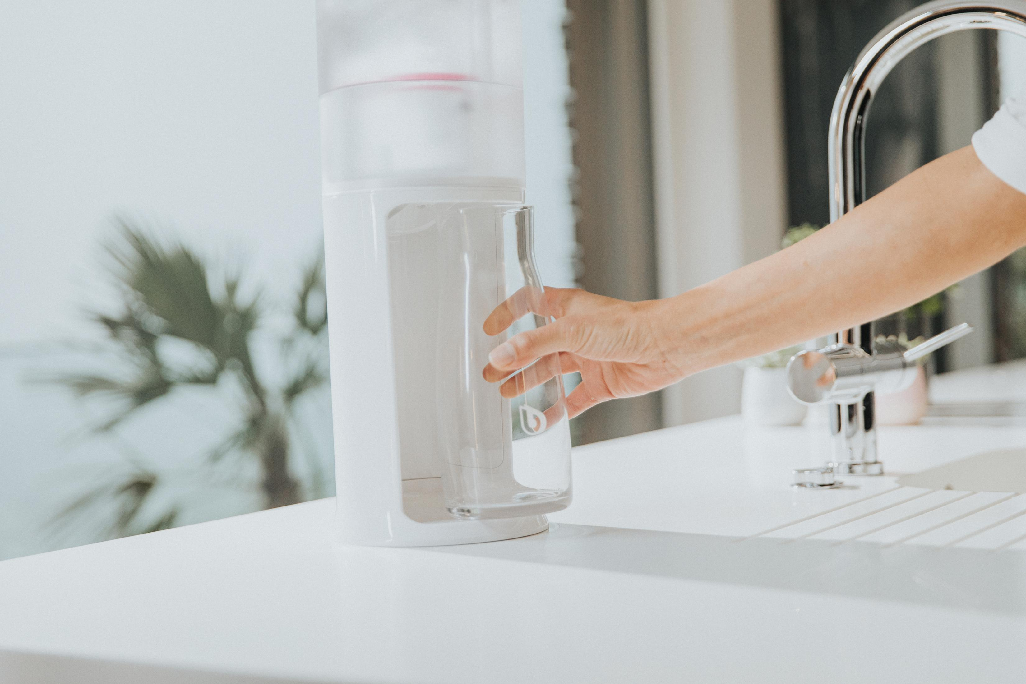 Eine Hand greift nach einem Glas. Ein weißer Wasserspender steht auf einer Arbeitsplatte neben einem Wasserhahn.