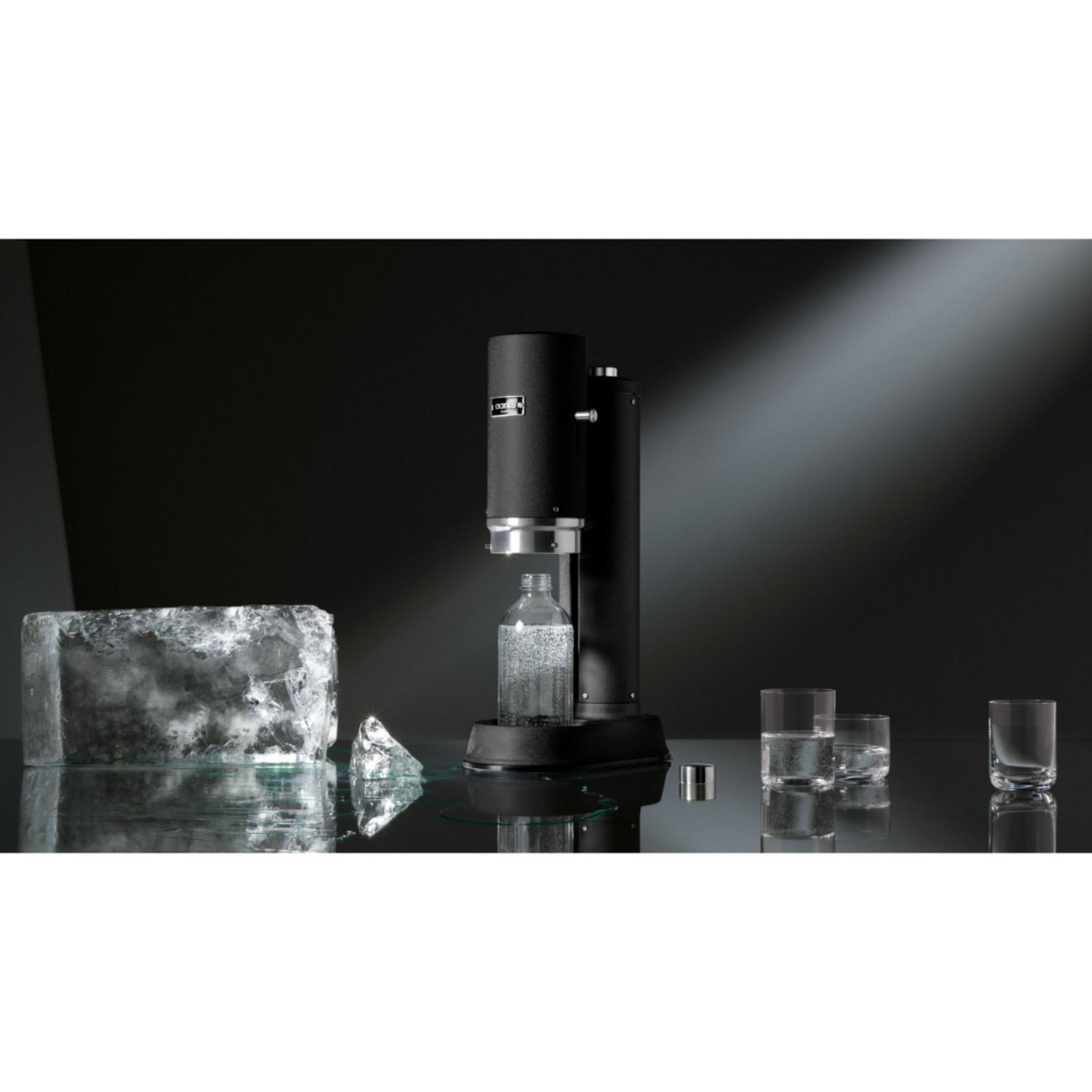 Soda maker nero con bottiglia di vetro e cubetti di ghiaccio su superficie riflettente.