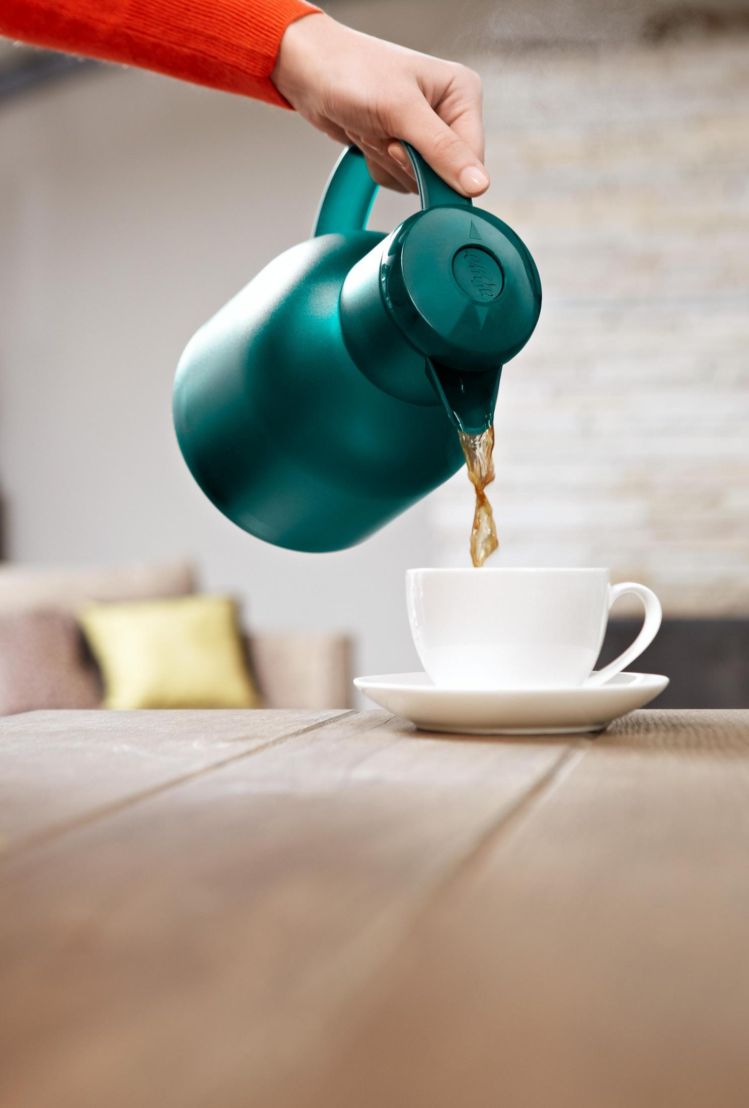 Eine Hand gießt braune Flüssigkeit aus einer tealfarbenen Thermoskanne in eine weiße Tasse.