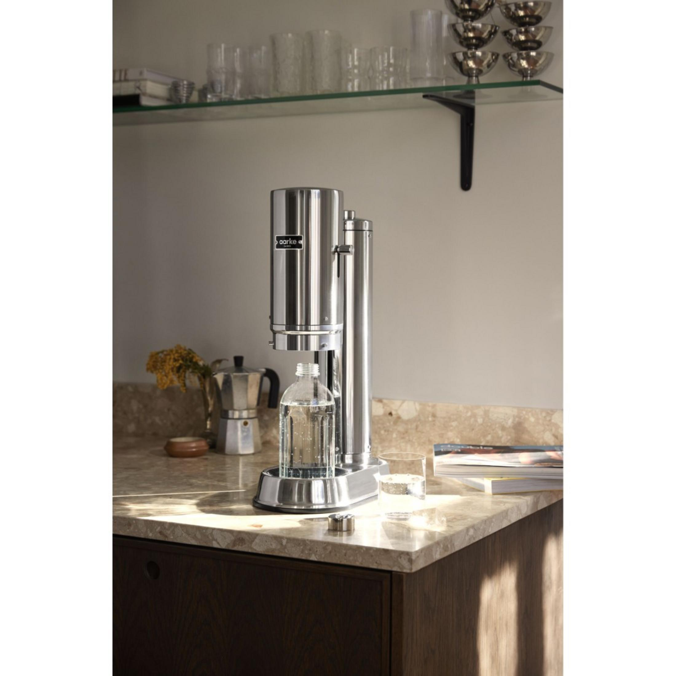 Silberner Soda-Maker mit Glasflasche auf Marmortisch, Kaffeemaschine und Buch.