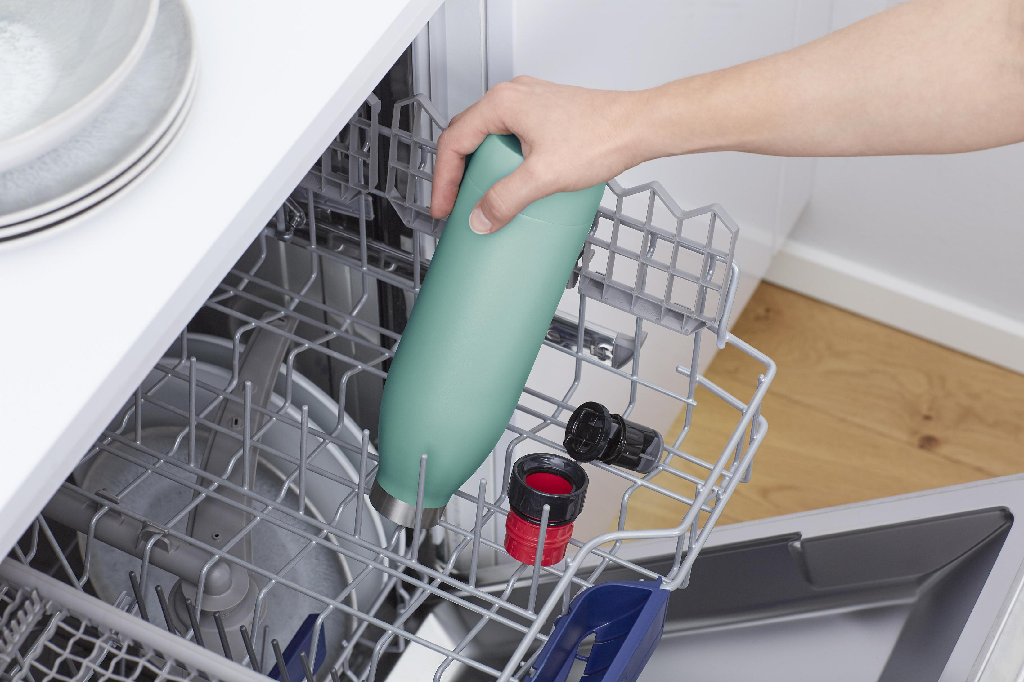 Eine Hand, die eine grüne Wasserflasche in einen Geschirrspüler legt. Andere Gegenstände sind sichtbar.