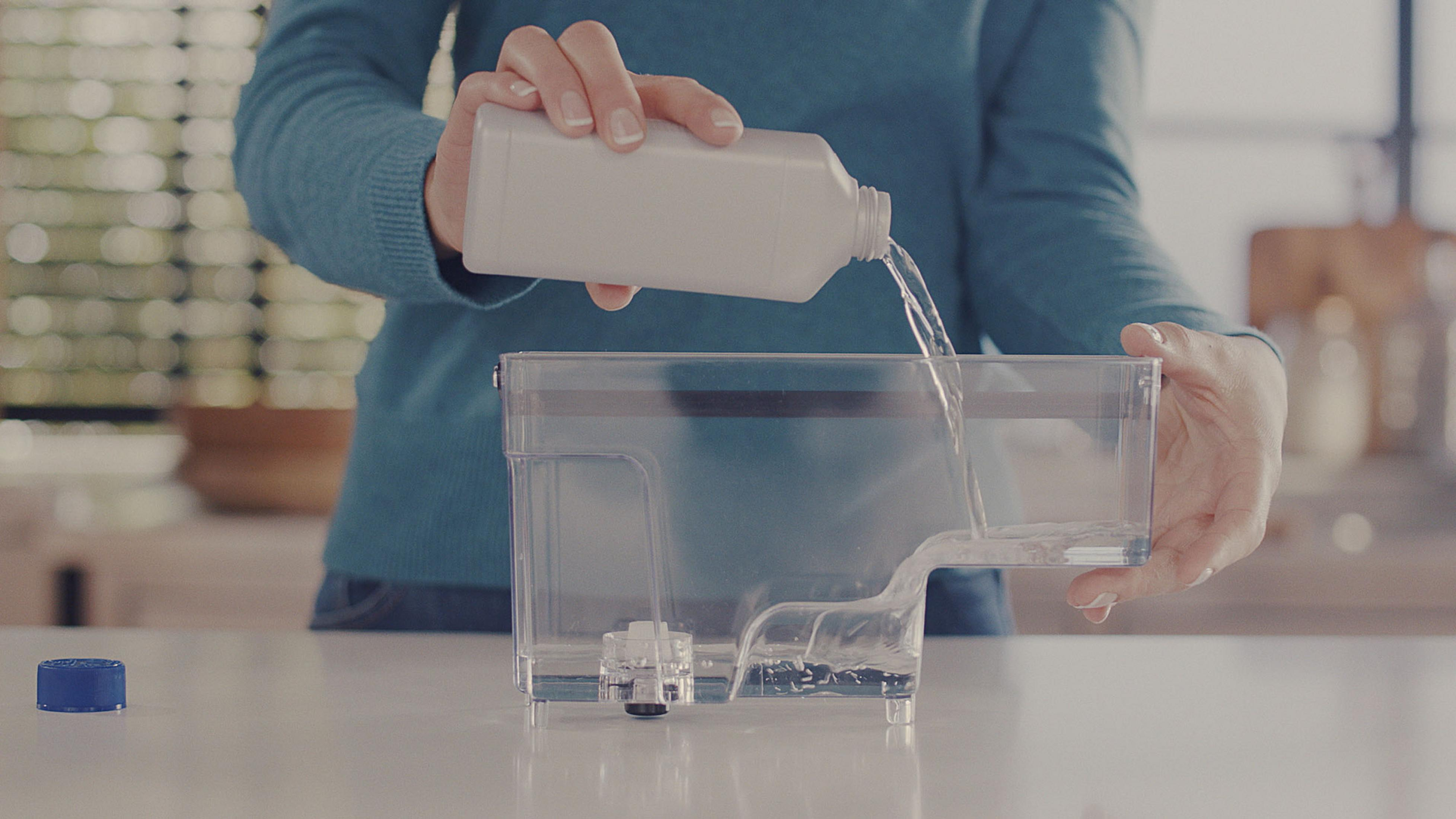 Person gießt Wasser in einen transparenten Wassertank. Weiße Theke, hellblaues Hemd.