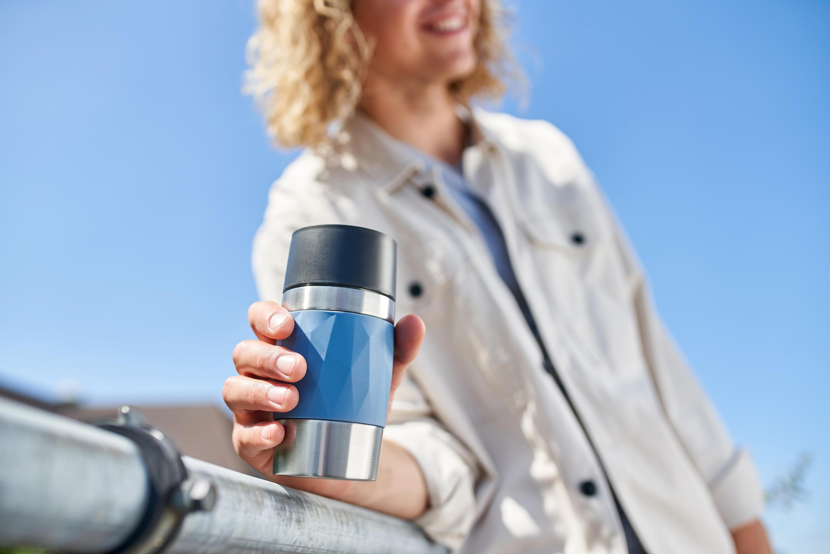 Eine Person hält einen blau-silbernen Reisebecher vor blauem Himmel. Sie lächelt.