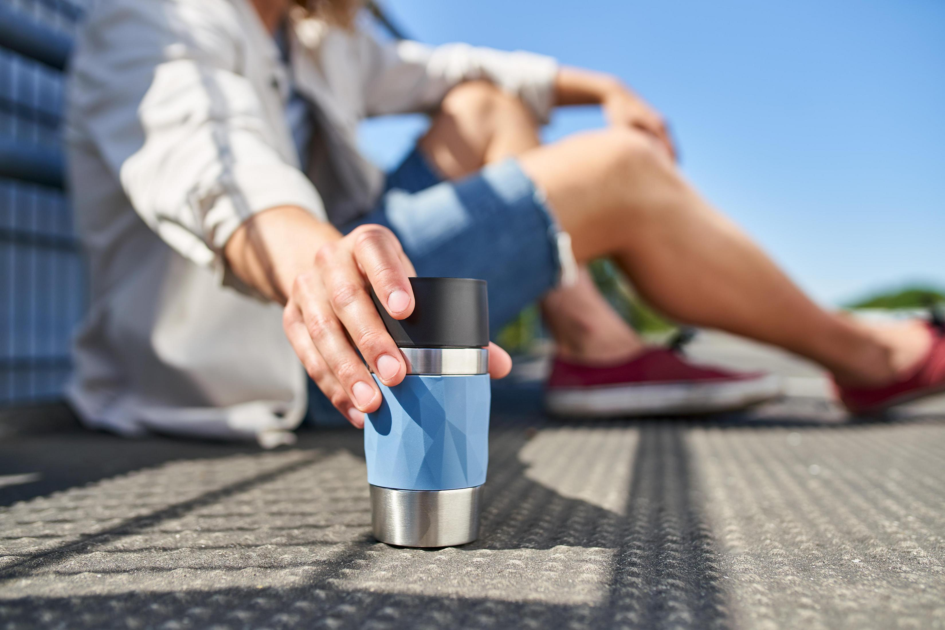 Person hält einen blau-silbernen Reisebecher. Draußen sitzen, sonniger Tag.