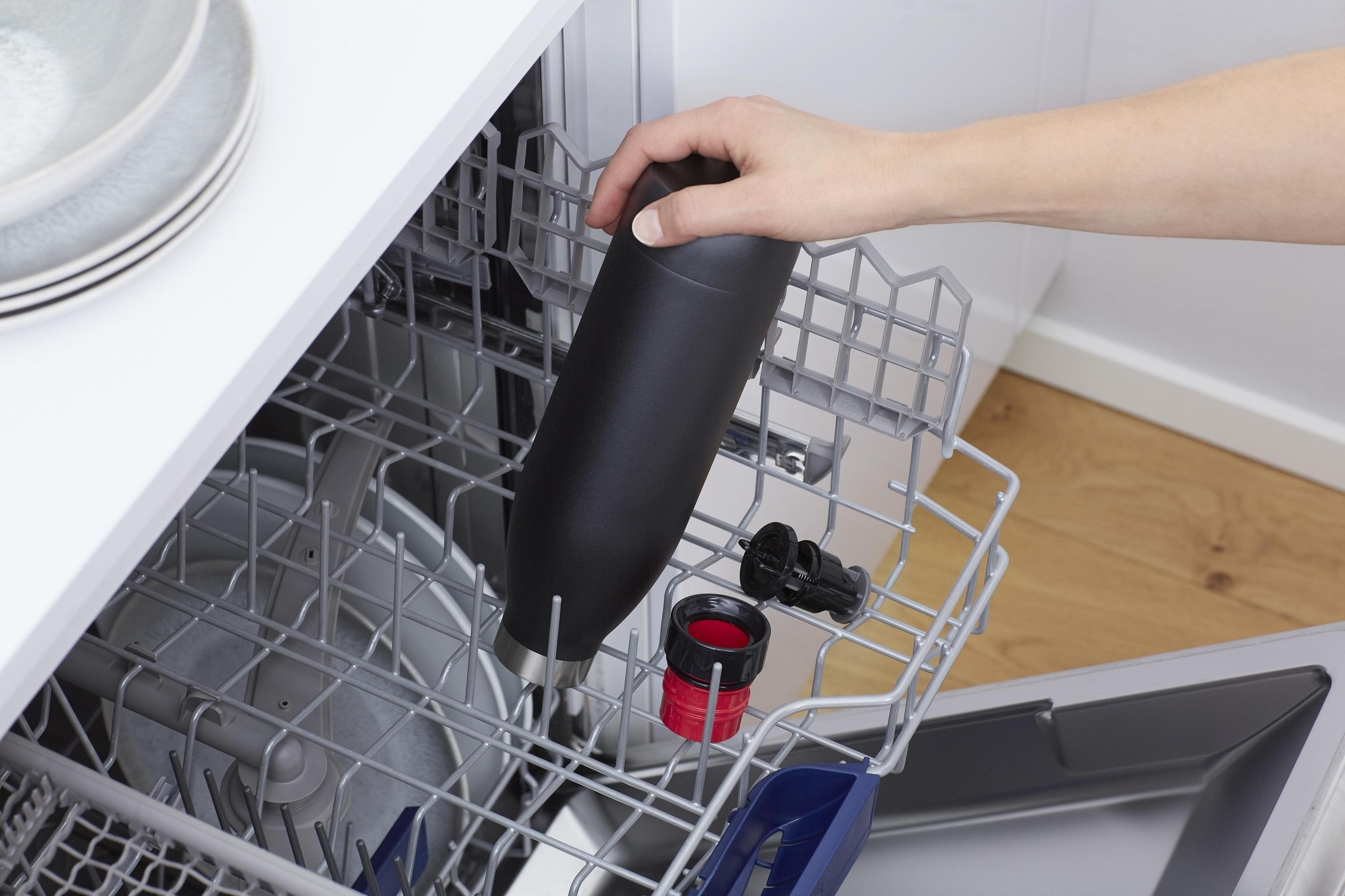Eine Person legt eine schwarze Wasserflasche in einen Geschirrspüler. Andere Gegenstände in der Maschine.