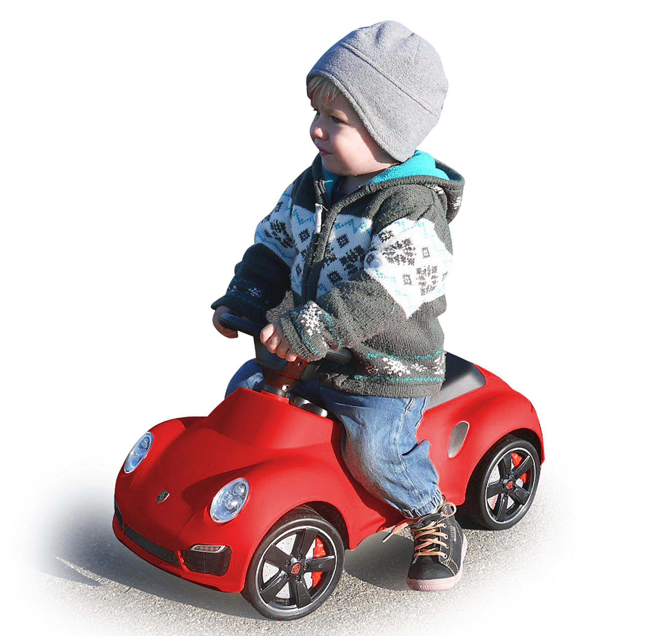 Un niño pequeño con gorro y chaqueta monta un coche de juguete rojo sobre fondo blanco.