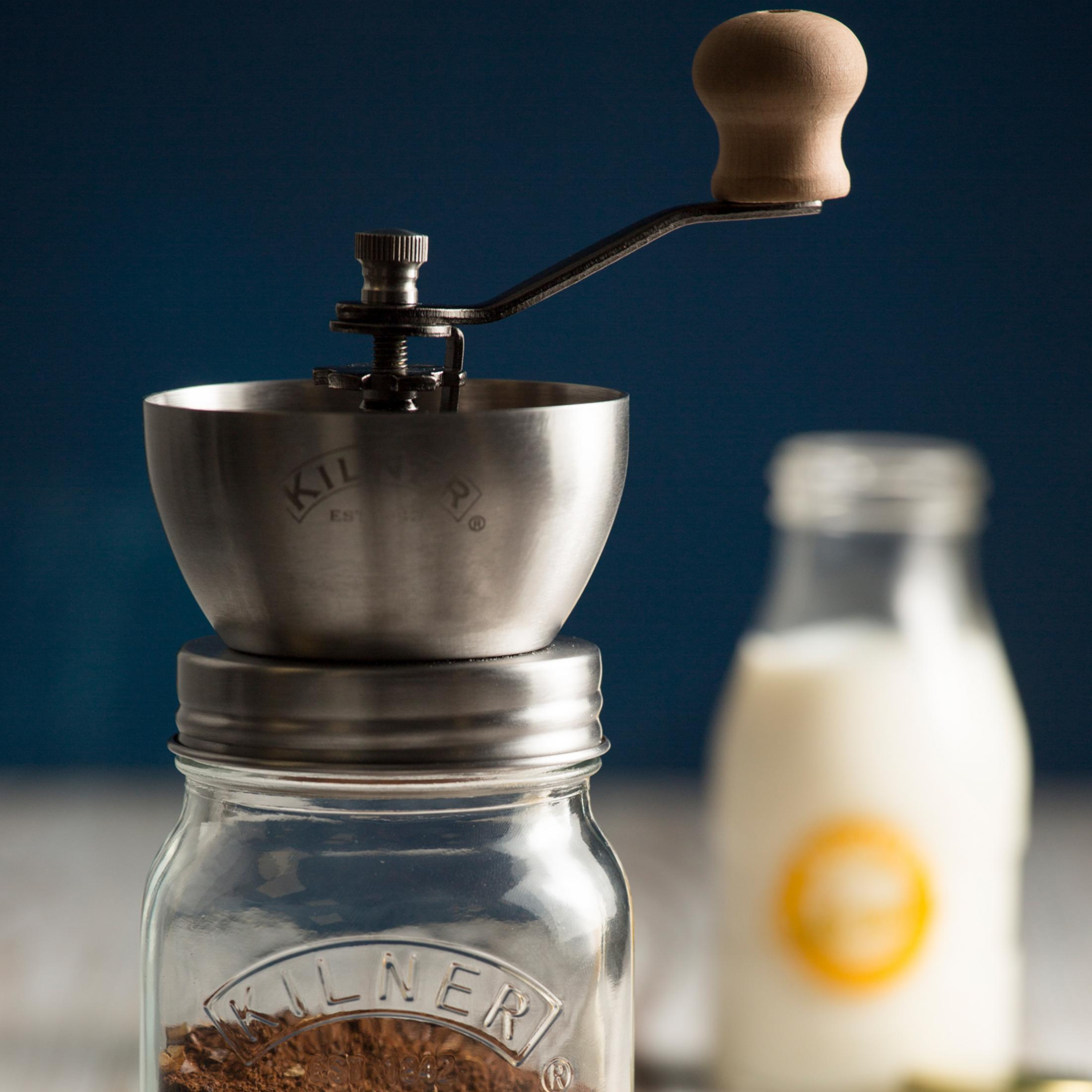 Eine Kaffeemühle auf einem Glas mit Kaffeepulver, im Hintergrund Milch in einer Flasche.
