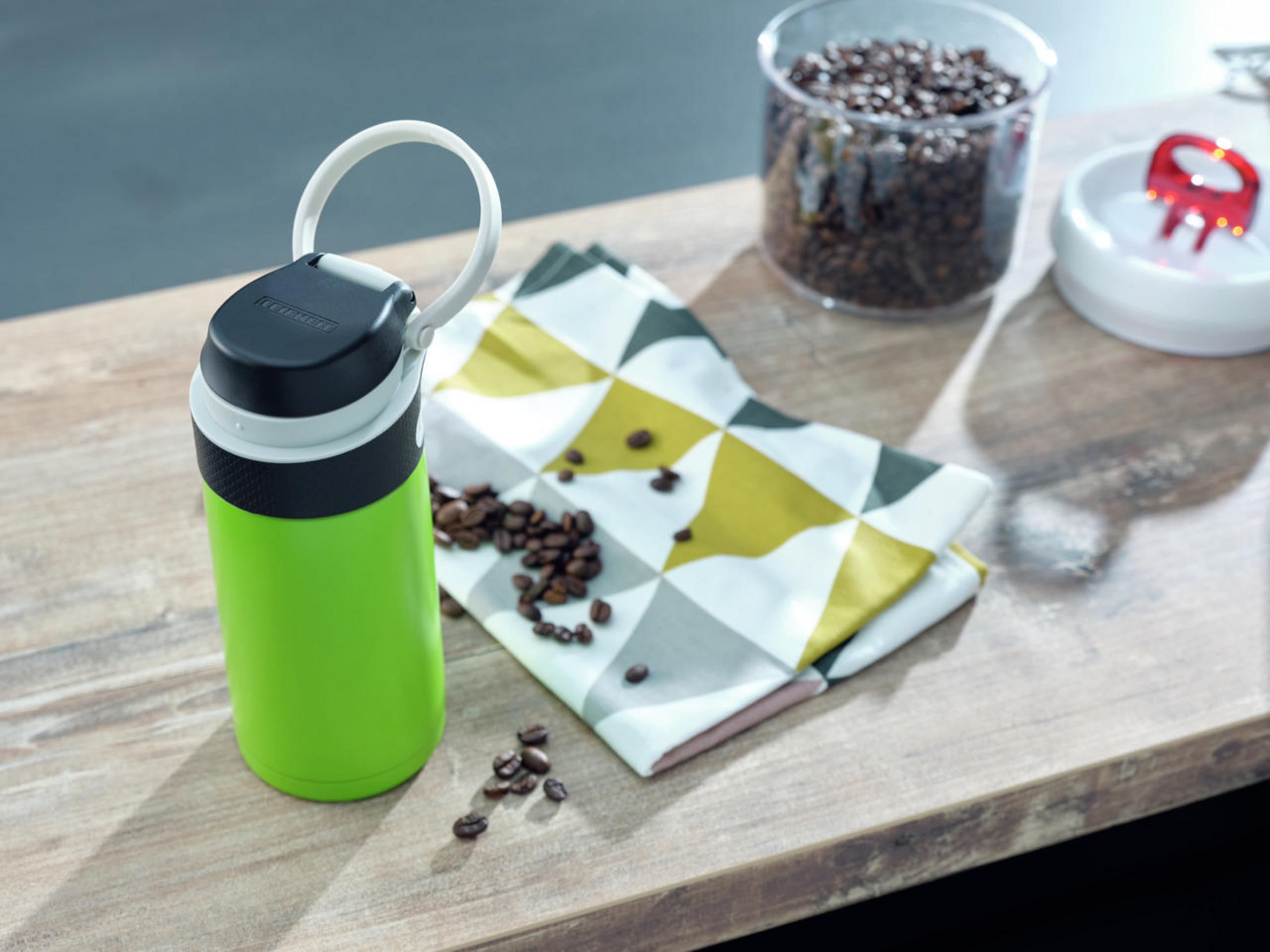 Eine grüne Flasche mit schwarzem Deckel und Griff, neben Kaffeebohnen und einer Serviette.