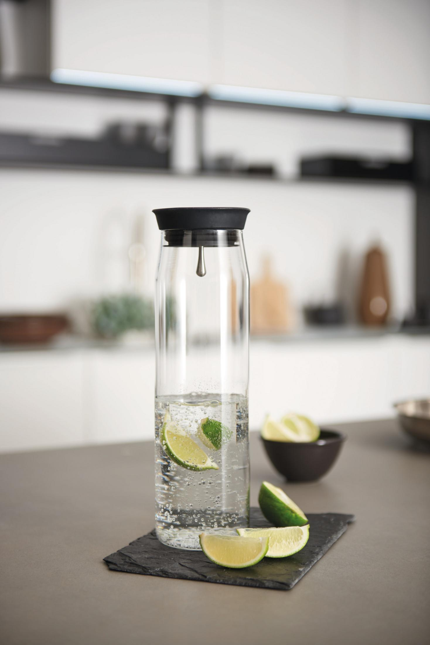 Hohe Glaswasserflasche mit Limettenscheiben auf Schiefer, in Küchenumgebung.