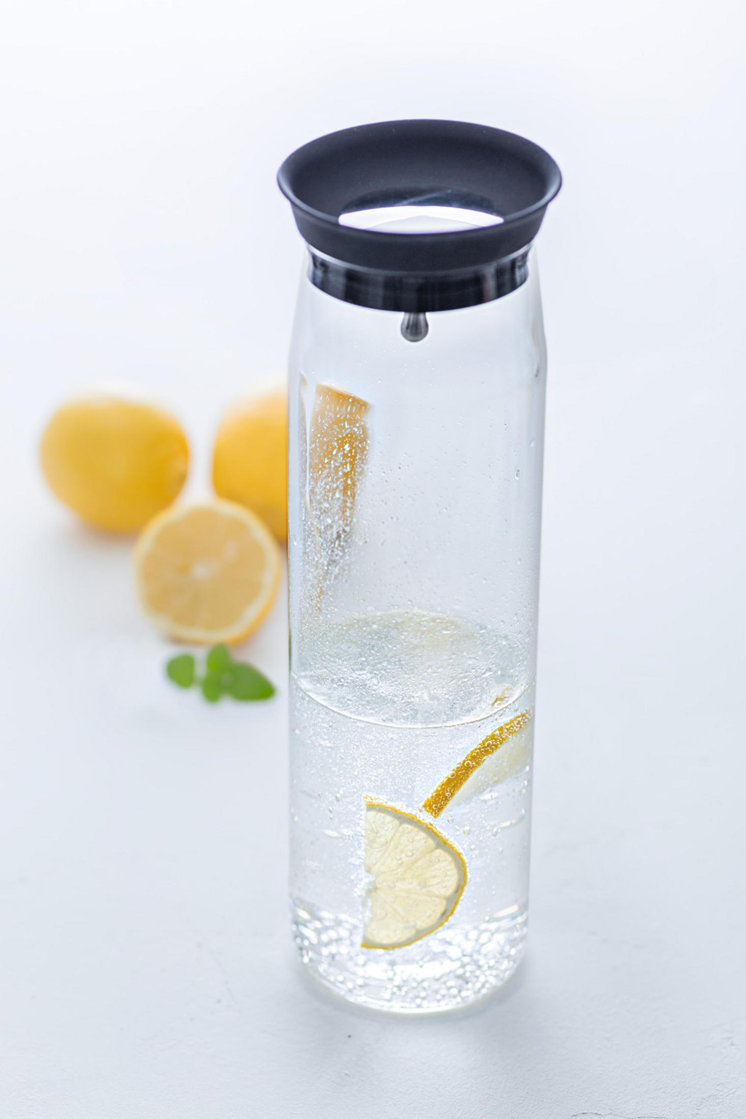 Eine klare Glaskaraffe mit Wasser, Zitronenscheiben. Zitronen im Hintergrund. Weiße Oberfläche.