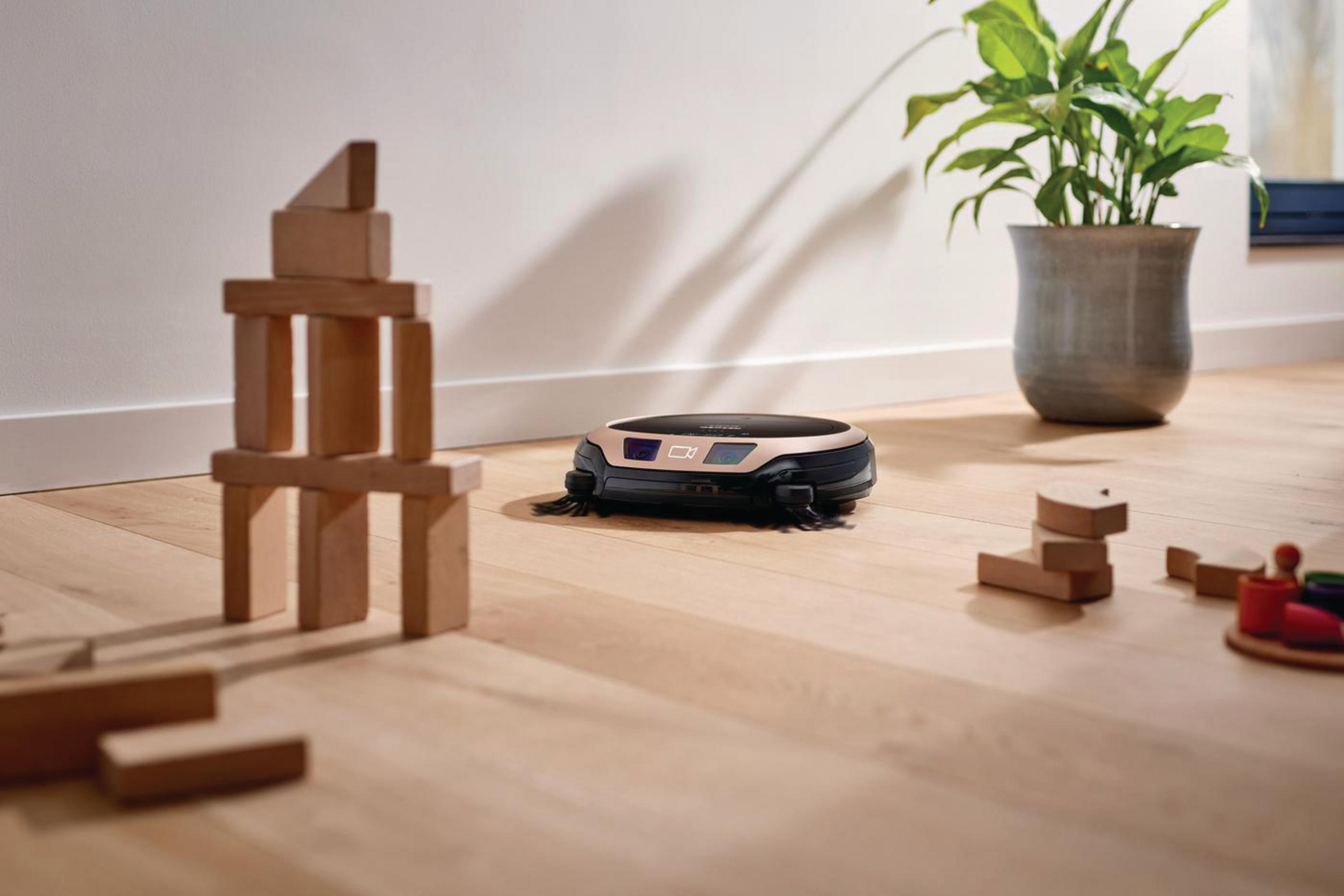 Ein Roboter-Staubsauger auf einem Holzboden. Holzblöcke und eine Pflanze im Hintergrund.