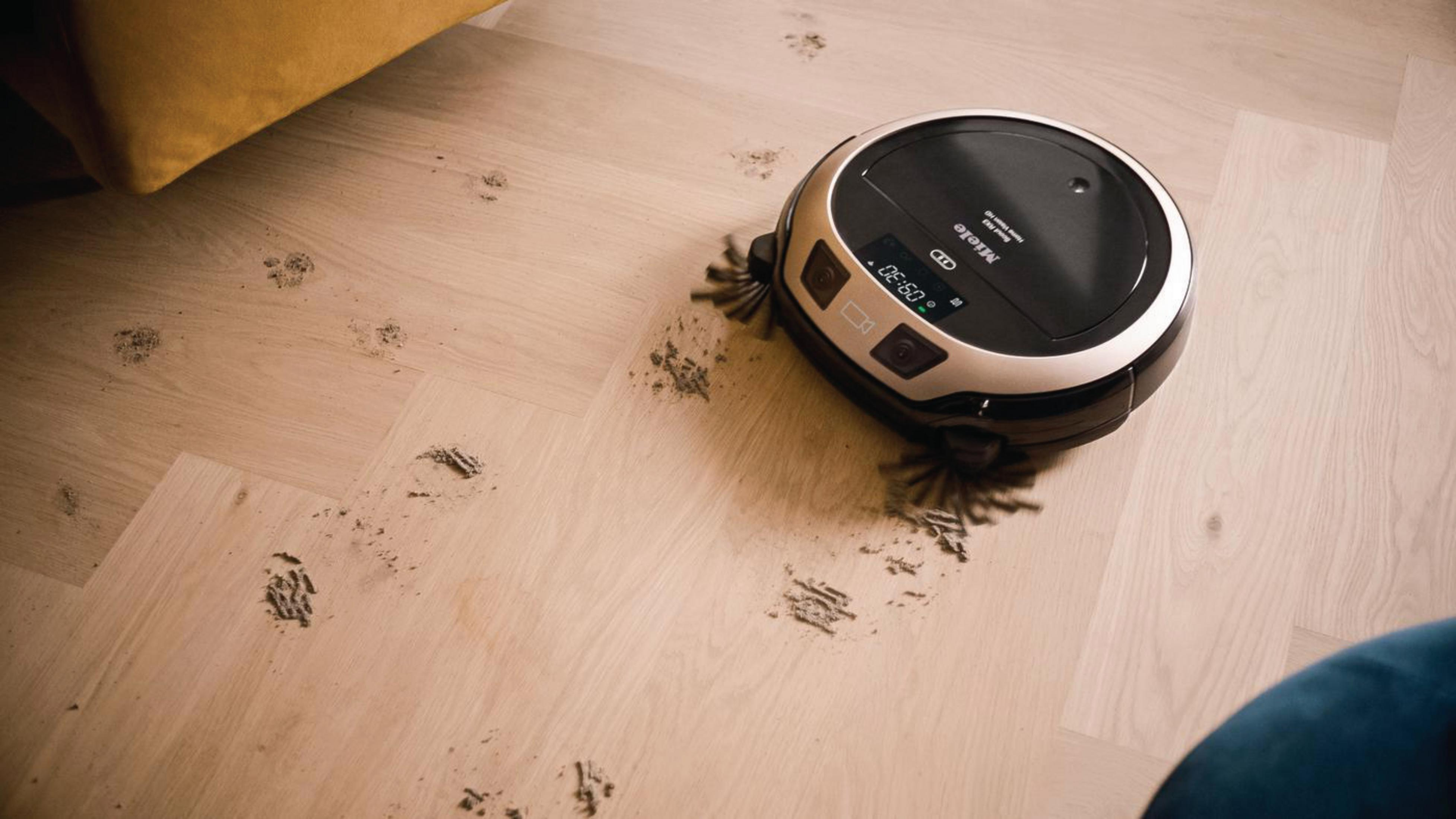 Roboter-Staubsauger auf Holzboden. Der Boden hat Schmutzspuren und blauen Stuhl sichtbar.